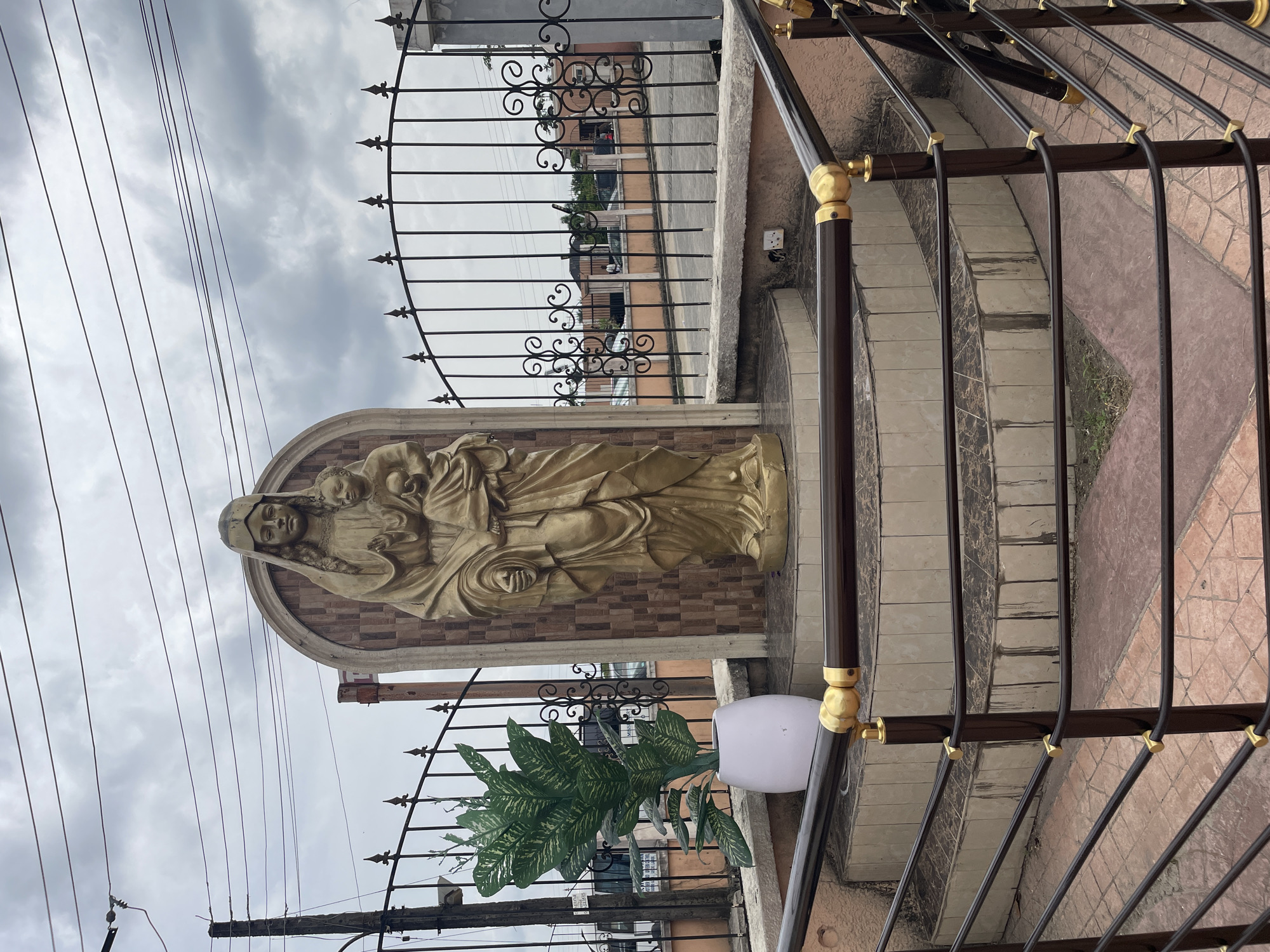 A vertical outdoor shot depicts a golden statue of a woman holding a child, mounted on a tiled wall, possibly representing the Virgin Mary and the infant Jesus. The statue is positioned near a metal fence with ornate designs, in front of a building. To the right of the statue, there's a stairway with metal railings that have decorative gold accents. A potted plant sits near the base of the statue, adding a touch of greenery to the scene. Overhead, power lines crisscross against a cloudy sky. The location appears to be an urban setting, given the presence of buildings and infrastructure, but the exact city or region is unknown.