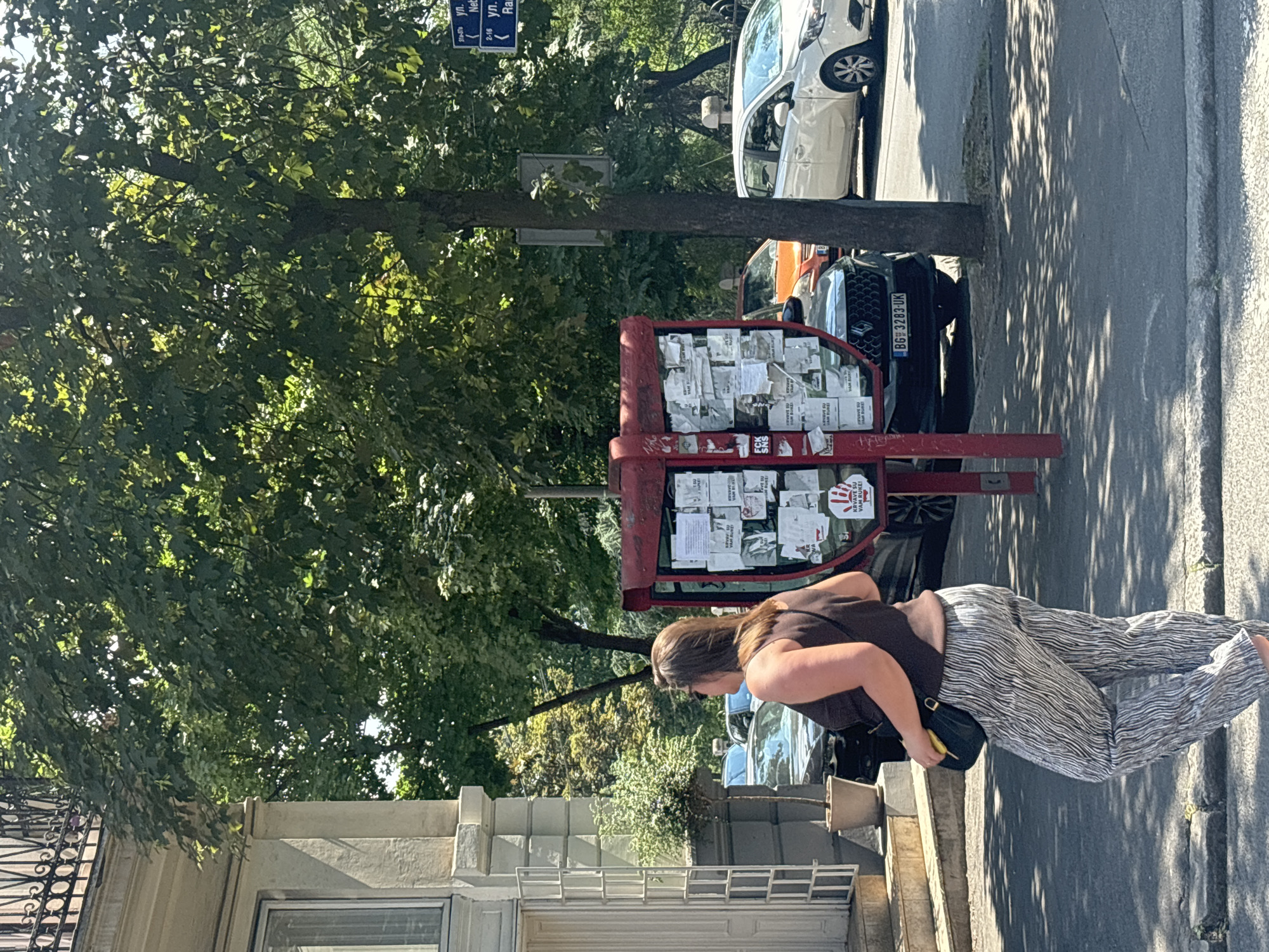 A woman in a brown tank top and patterned pants walks past a red bulletin board filled with papers. She is holding a black purse and a yellow object, possibly a fruit, in her right hand. The bulletin board is positioned on a sidewalk next to a street with cars parked on it. Behind the bulletin board, trees with green leaves create dappled shade on the ground. A white car is visible in the background, along with another dark-colored car. A building with architectural details is on the left side of the image. A blue street sign is visible in the upper left corner. The scene appears to be set outdoors during the daytime, likely on a sunny day given the shadows. The overall atmosphere seems casual and urban. The license plate of the dark car reads "BG 3283-UK". Some of the text on the bulletin board is in Cyrillic script.
