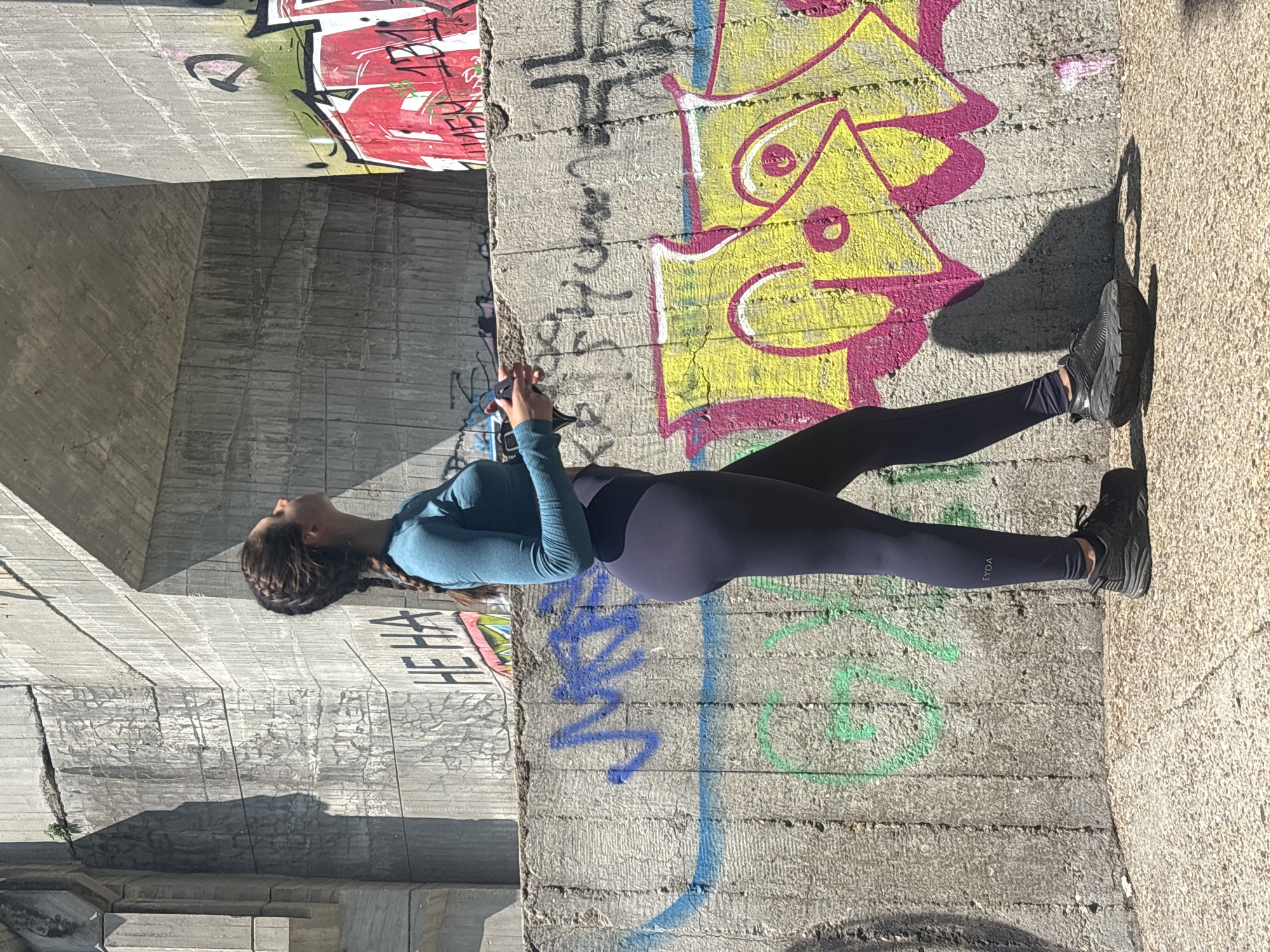 The image captures a young adult female in profile, facing left and looking upward, standing against a backdrop of heavily graffitied concrete structures under bright daylight in Varna, Bulgaria. She has dark hair styled in two braids and is wearing a long-sleeved, fitted teal top, dark navy leggings with the "EYDA" logo visible on her left calf, and black athletic shoes with thick soles. Her hands are clasped at chest height, holding a dark-colored smartphone. The background consists of large, textured concrete walls and an overhead concrete structure, indicative of an overpass or bridge. These concrete surfaces are extensively covered in various graffiti, including the legible text "HE HA" on an upper wall, a prominent red and yellow piece with abstract shapes, and a black hammer and sickle symbol outlined in red on the upper right wall. The ground is a light-colored, textured pavement. Intense sunlight from the right casts sharp, distinct shadows of the structures and the female onto the surrounding surfaces.