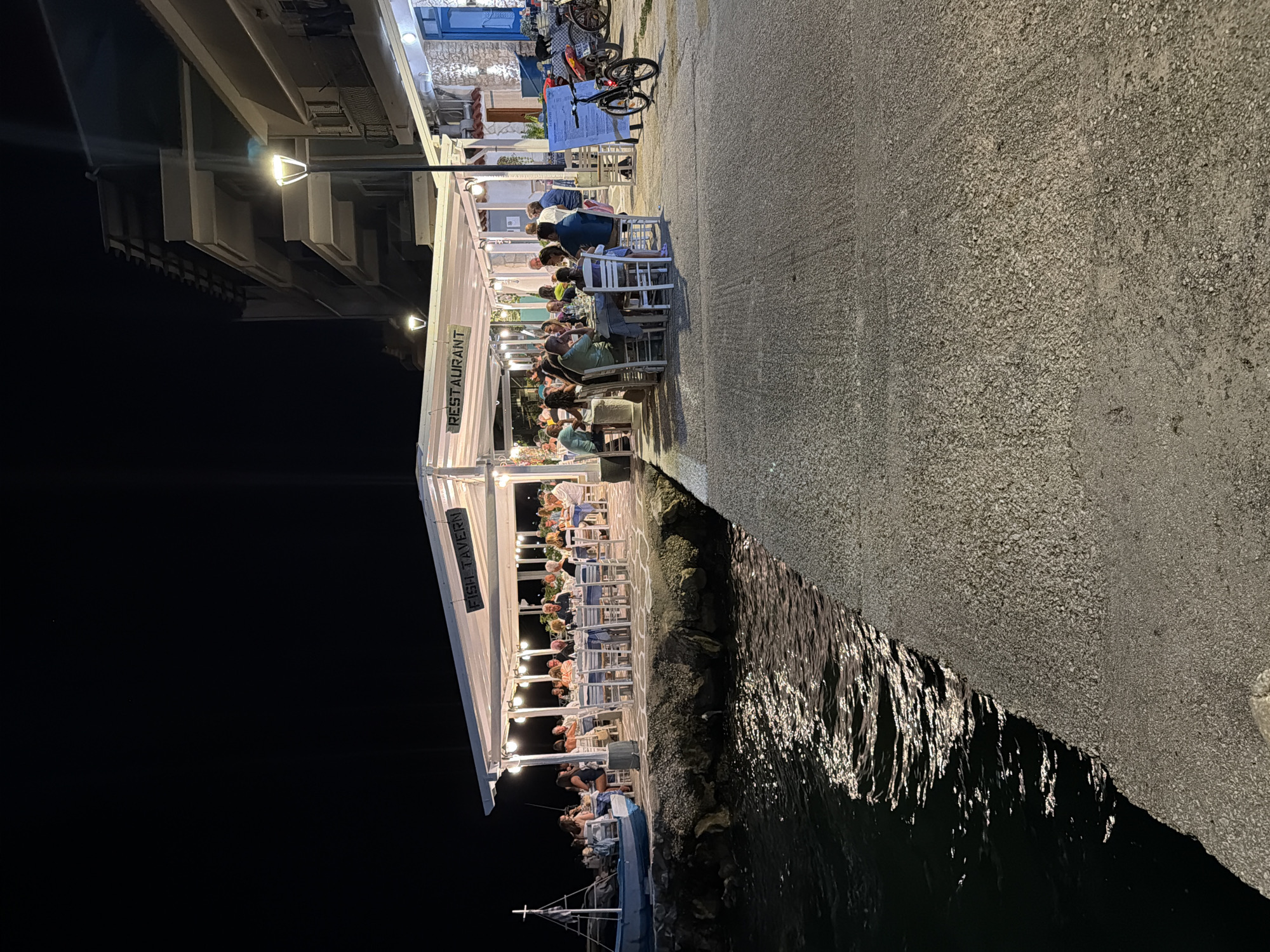 A night scene in the Municipal Unit of Alyzia, Greece, shows an outdoor restaurant situated on a waterfront. A covered seating area with white tables and chairs is filled with patrons dining. A sign above the covered area reads "FISH TAVERN RESTAURANT." The restaurant is illuminated by string lights. A small boat with a flag is docked to the left of the restaurant. The water in front of the restaurant reflects the lights. A paved walkway extends from the right of the image towards the restaurant. A bicycle is parked near a sign on the right side of the walkway. A building with balconies is visible behind the restaurant. The sky is dark, indicating it is nighttime.