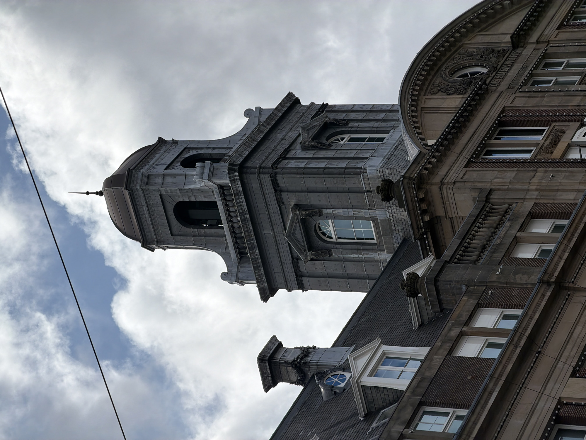 The image captures the top section of an ornate stone building, likely a historical edifice, under a cloudy sky. The building features a prominent bell tower with a domed roof topped by a spire. The tower is constructed of stone blocks and has arched openings. To the left of the tower, a sloped, dark-tiled roof is visible, with a decorative chimney stack and dormer windows. The lower portion of the building showcases classical architectural elements, including detailed carvings and a series of windows. The text "DE BUENKORE" is inscribed above a rounded section of the building's facade. A dark cable stretches across the upper portion of the image, indicating the presence of overhead wires. The scene appears to be an exterior urban environment in Amsterdam, Netherlands.
