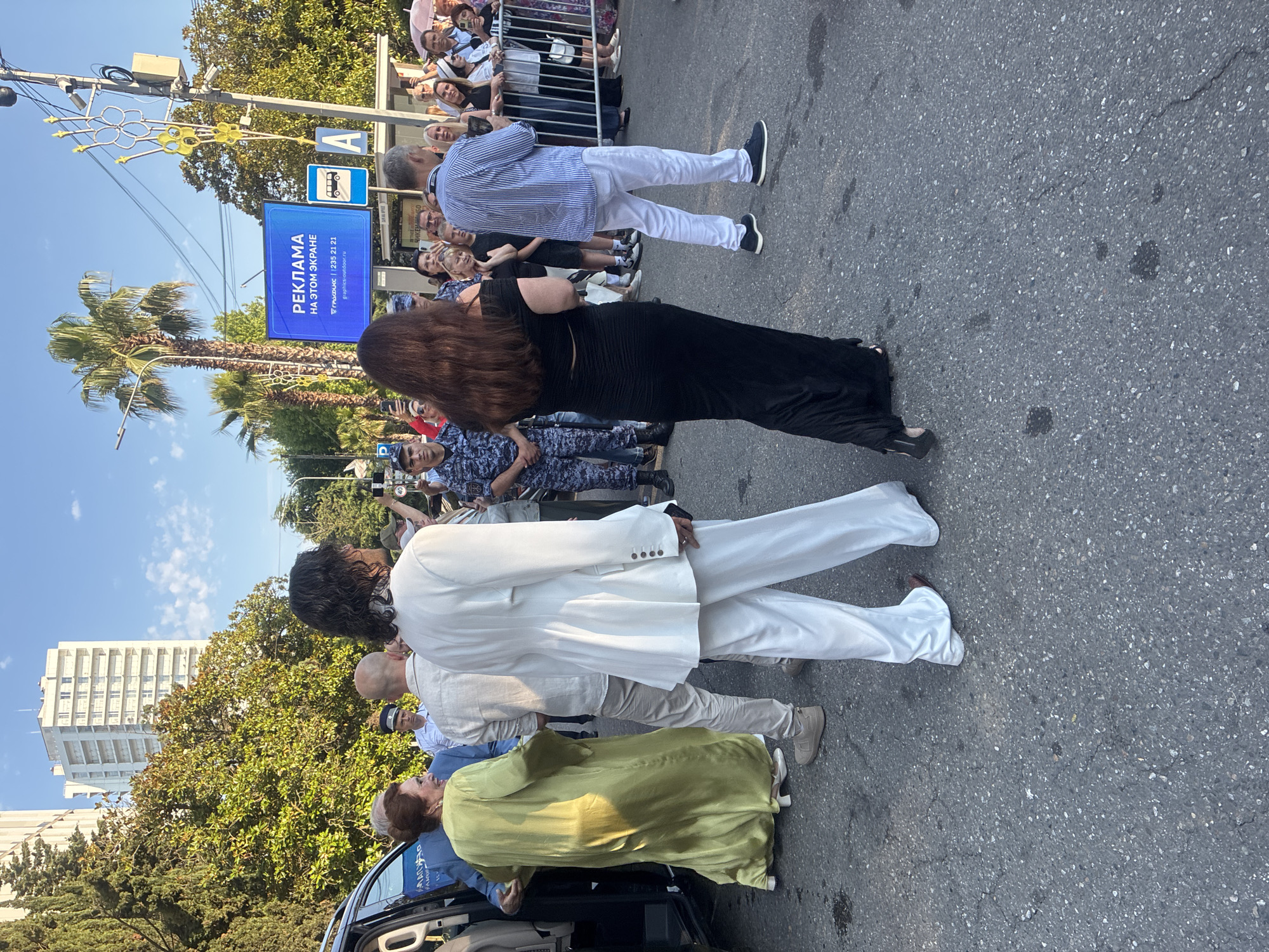 The image depicts a group of people outdoors on a street, possibly in Sochi, Russia. The scene is set on a sunny day, with a clear blue sky visible. A tall, modern white building and lush green trees are in the background.
The main subjects are several individuals, including a woman with long dark hair wearing a black dress, a man in a white suit and pants, and another person in a light-colored outfit. There are also people in the background wearing what appear to be police or security uniforms. A black car is partially visible at the bottom of the frame.
The people are gathered on a paved street, with some standing and others near the car. The overall atmosphere suggests a public event or gathering, possibly a formal occasion given the attire of some individuals. The presence of a crowd and security personnel hints at an organized event, perhaps a red carpet or arrival at a venue.
Visible text on a blue sign reads "РЕКЛАМА НА ЭТОМ ЭКРАНЕ" which translates to "ADVERTISEMENT ON THIS SCREEN." Below that, there is a phone number and a website address that appears to be "gruppatrax.ru." Another sign with a bus symbol is also visible. The time of day appears to be daytime, given the bright sunlight casting shadows.
