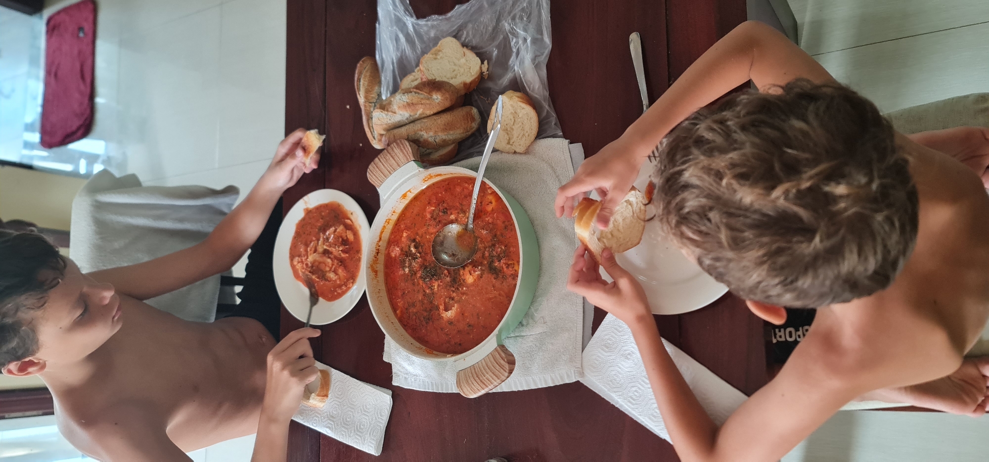 Two shirtless young boys are seated at a dark brown wooden table, eating a meal consisting of a red-sauce soup and bread. A large, light-green pot filled with the soup sits in the center of the table, along with a ladle. A plastic bag containing baguette-style bread is near the pot. Each boy has a white plate with a serving of the soup and a piece of bread. One boy on the left is eating with a fork and a piece of bread in his hands, while the other boy on the right is tearing a piece of bread. A white towel or napkin is placed under the pot and each plate. The location is likely indoors, with a glimpse of an air conditioner and a maroon cloth in the background. Considering the likely geographic location of the image as Đà Nẵng, Vietnam, it is likely that the food is a local dish that is eaten at home.