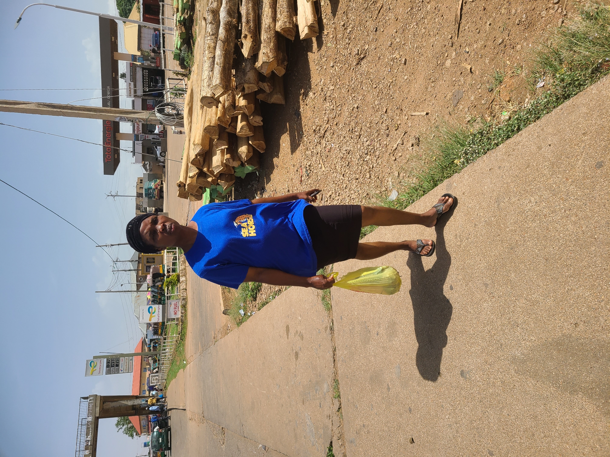 Here is a forensic description of the image:
**Scene:** The image captures a person standing on a sidewalk next to a pile of timber logs. The location appears to be an urban street, evidenced by commercial establishments and infrastructure in the background.
**Subject:** A person is positioned in the foreground wearing a blue T-shirt, black shorts, and sandals. They are holding a yellow plastic bag.
**Objects:** A stack of timber logs lies adjacent to the sidewalk. A gas station is visible in the background. Additionally, there's urban street infrastructure, including utility poles, signage, and a pedestrian overpass.
**Actions and Interactions:** The person is standing stationary and appears to be posing for the image.
**Environment:** The scene is set during daylight hours with a blue sky. The environment reflects an urban landscape with a mix of commercial and possibly residential elements.
**Location:** There's signage for "TotalEnergies" and "Total Energie" which can help identifying the city and region. The signboard mentioning "KUBU GOWON" is also helpful for location identification.