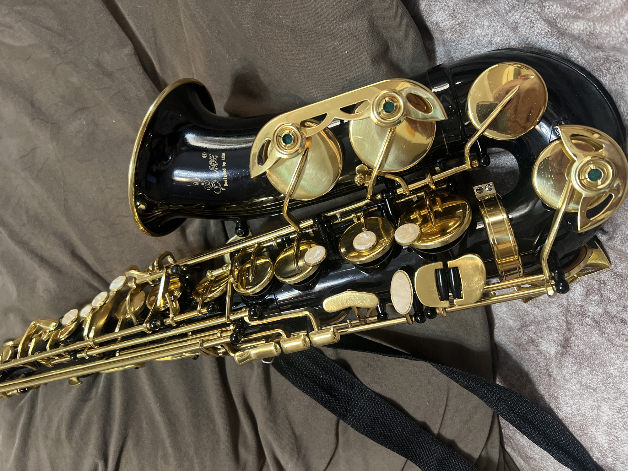 A black saxophone with gold accents rests on a brown, textured surface. The saxophone is angled, with its bell facing towards the bottom right of the frame. Several keys and pads are visible along the instrument's body. A black strap is attached to the saxophone and hangs down the left side. The background is a soft, wrinkled brown fabric. There is no indication of location in the city of Masazir, Azerbaijan within the image.