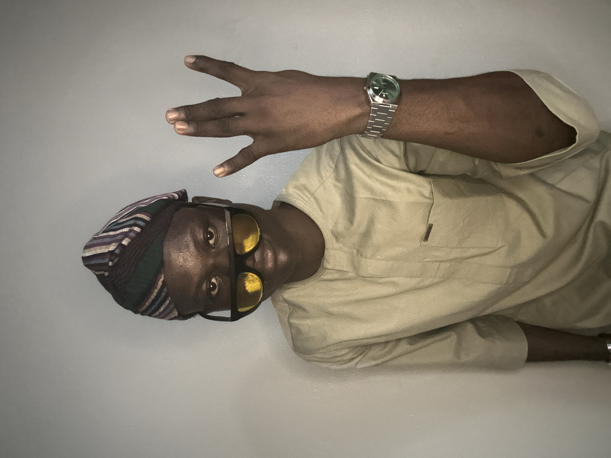 This is a vertically oriented, full-body studio portrait of a young Black man wearing a beige dashiki and a colorful striped cap. He is also wearing sunglasses with yellow lenses and a silver wristwatch on his left wrist. His right hand is raised, with his index and middle fingers extended and his thumb, ring, and pinky fingers curled. The background is a plain, light gray wall. The lighting suggests an indoor setting, likely with artificial light, and there are no indications of the time of day or weather. The overall mood is casual and confident. The man is facing the camera, with a slight smile. The clothing and cap suggest a possible cultural context related to West Africa, specifically Nigeria, given the location context. No text is visible in the image.
