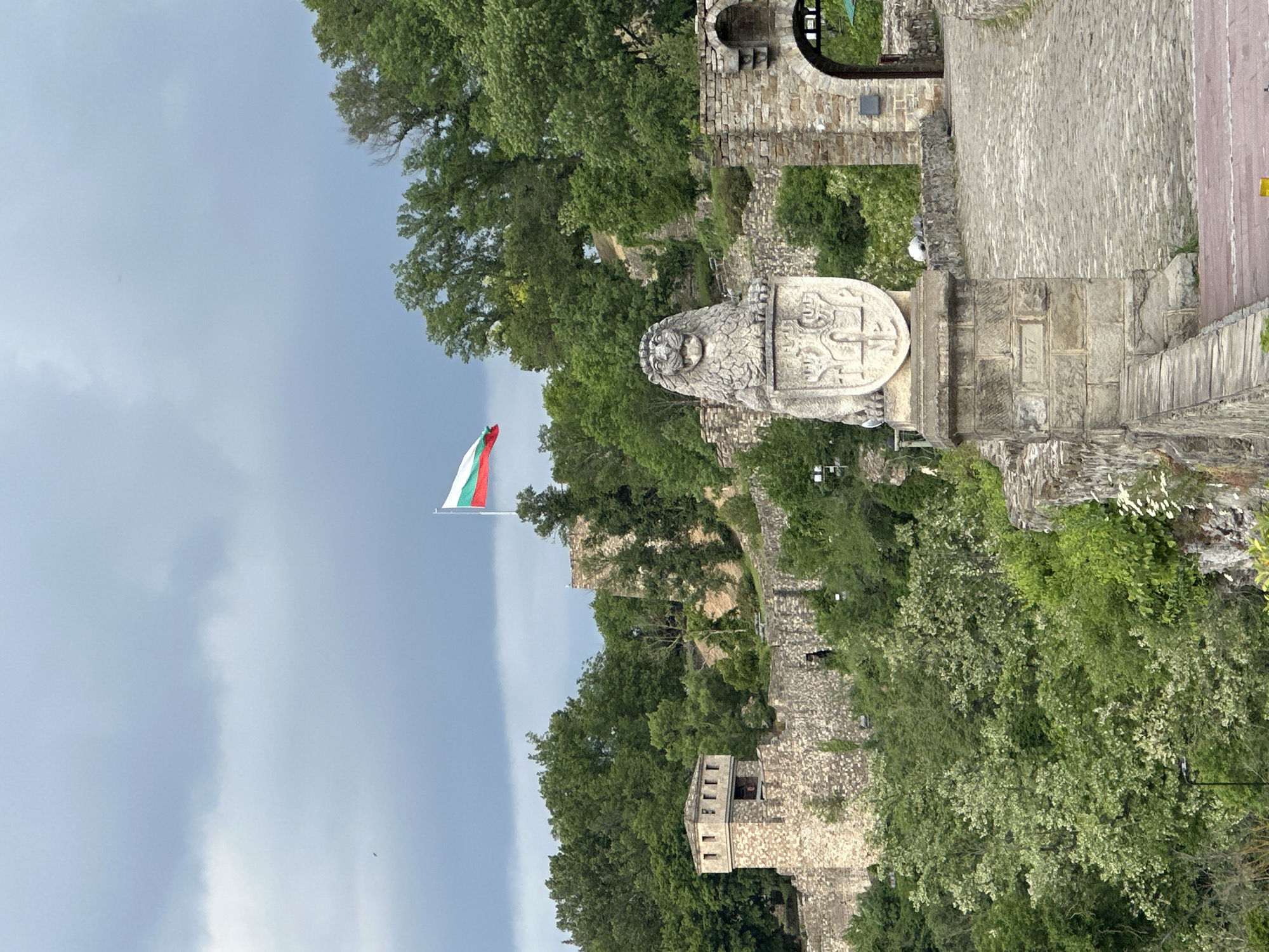 The image shows a view of Tsarevets Fortress in Veliko Tarnovo, Bulgaria, with a Bulgarian flag flying. The fortress is built on a hill and features stone walls, towers, and gate structures surrounded by lush green trees. A stone lion statue stands on a bridge or wall, likely an emblem or memorial within the fortress. The sky is overcast, suggesting cloudy weather.