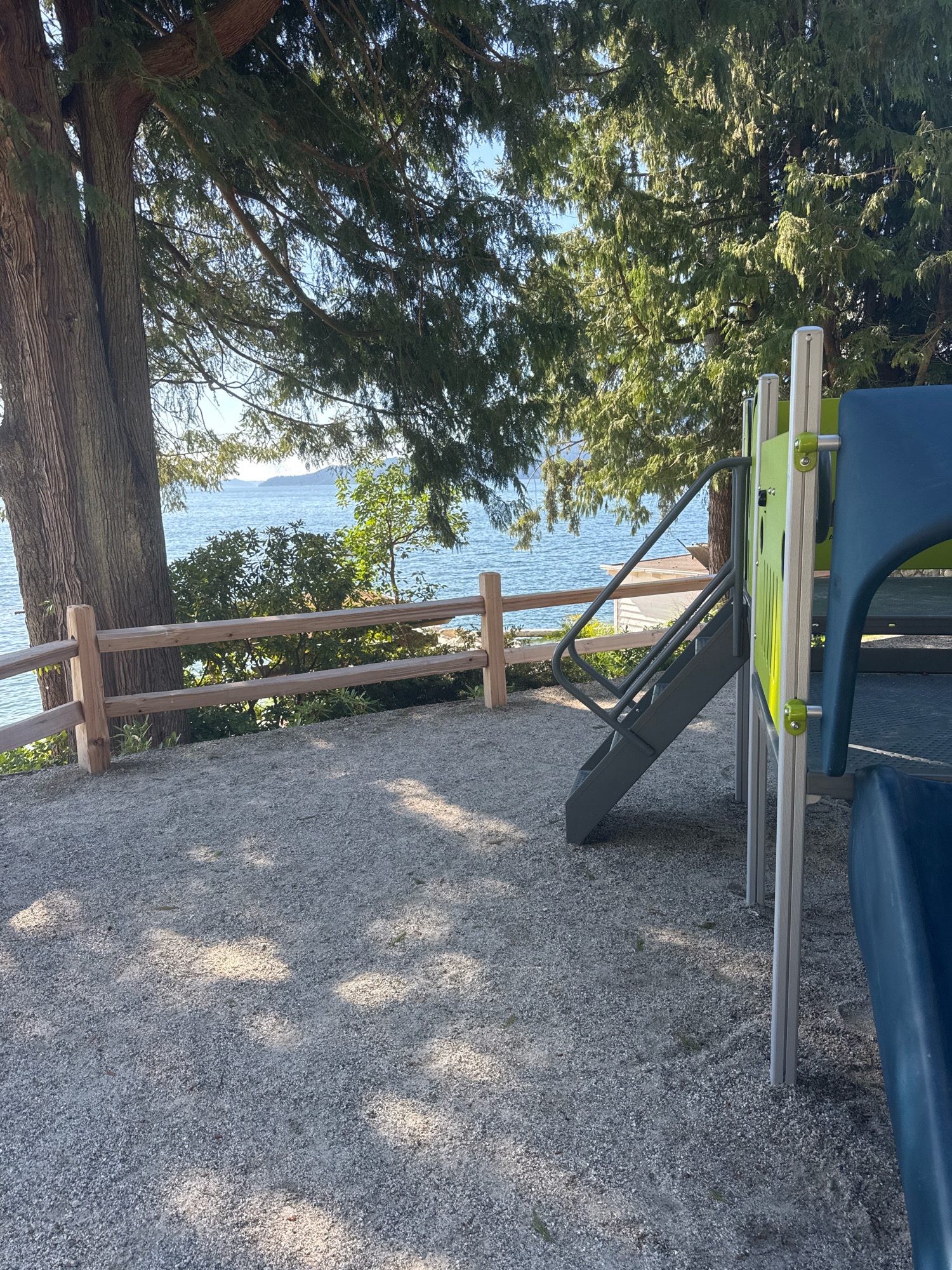 In the Canadian city of Lions Bay, a playground overlooks a body of water. A thick tree trunk and evergreen branches dominate the left side of the frame, with sunlight dappling through the foliage onto a gravel ground cover. A wooden fence runs horizontally across the midground, separating the playground area from a lush green shrubbery. Beyond the fence, the blue water of the ocean is visible, with faint landmasses in the distance under a clear sky. A grey metal staircase, part of a playground structure, ascends towards a blue and green plastic slide on the right. The playground equipment is modern and appears to be made of metal and plastic. The scene is tranquil and bathed in natural light.