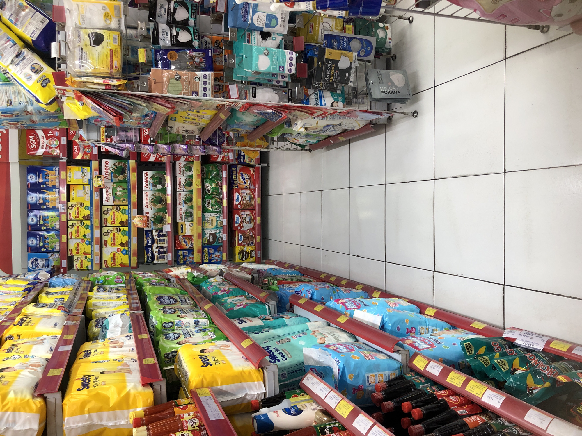 The image shows a retail aisle in a convenience store, likely in Putat, Indonesia, judging by the Indonesian product labels and store signage. The aisle is lined with shelves stocked with various consumer goods. On the left, shelves display bottles of sauces and condiments, including what appears to be soy sauce and chili sauce. Above these, there are packages of baby diapers and formula. The right side of the aisle features shelves with hygiene products, prominently displaying an assortment of face masks in various brands and packaging. Other items visible include baby wipes, and possibly some snacks or processed foods. The floor is tiled with white squares. The lighting appears to be fluorescent, typical of an indoor retail environment. There are no people visible in the image.