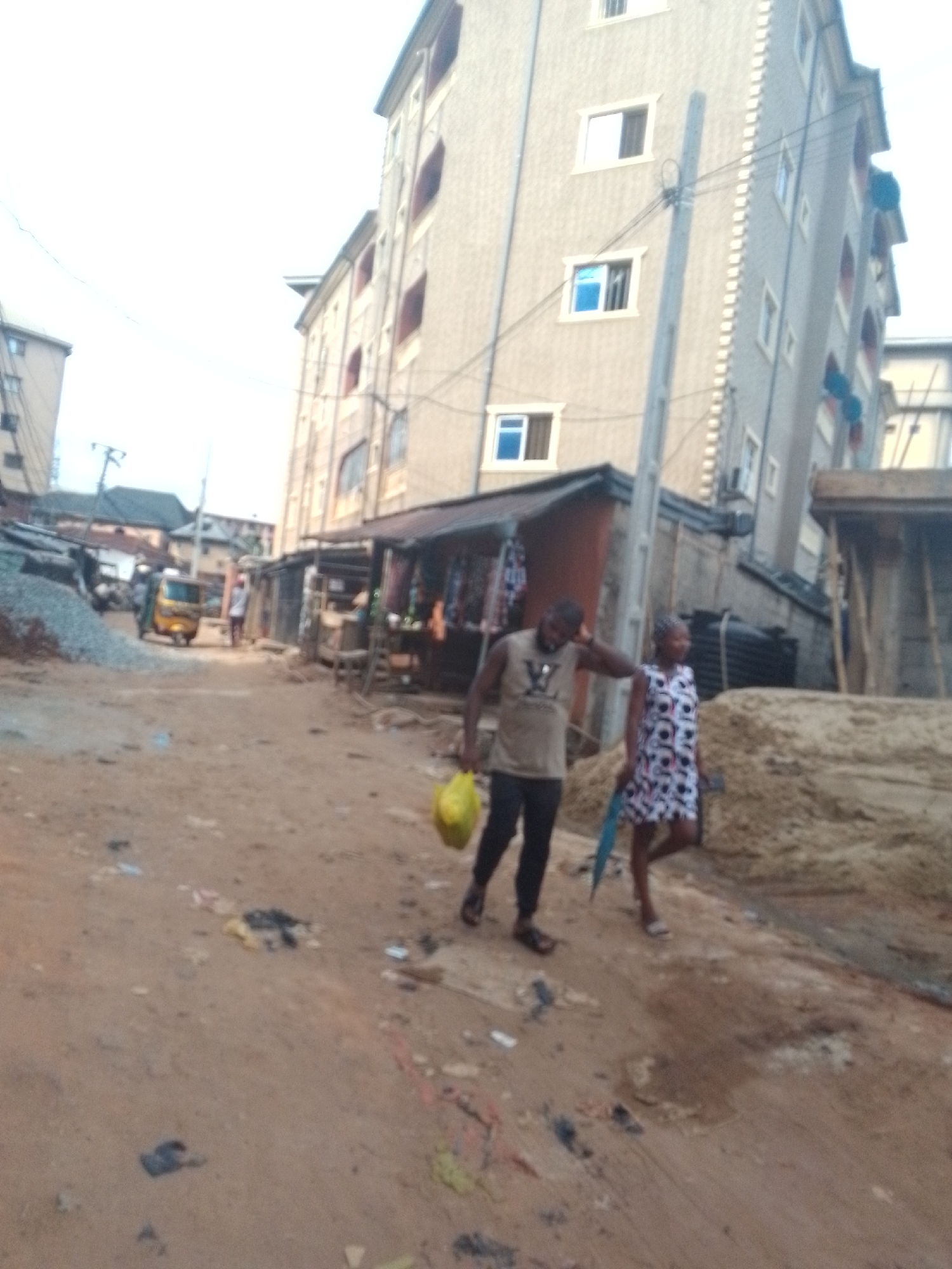 The image shows a street scene in Obosi, Nigeria. The street is unpaved and appears to be littered with debris. Two people, a man and a woman, are walking on the street. The man is wearing a tank top, dark pants, and sandals, and he is carrying a yellow bag. The woman is wearing a patterned dress and sandals. A motorcycle-taxi is driving down the street in the background. A large building rises behind the people and shops in the foreground.