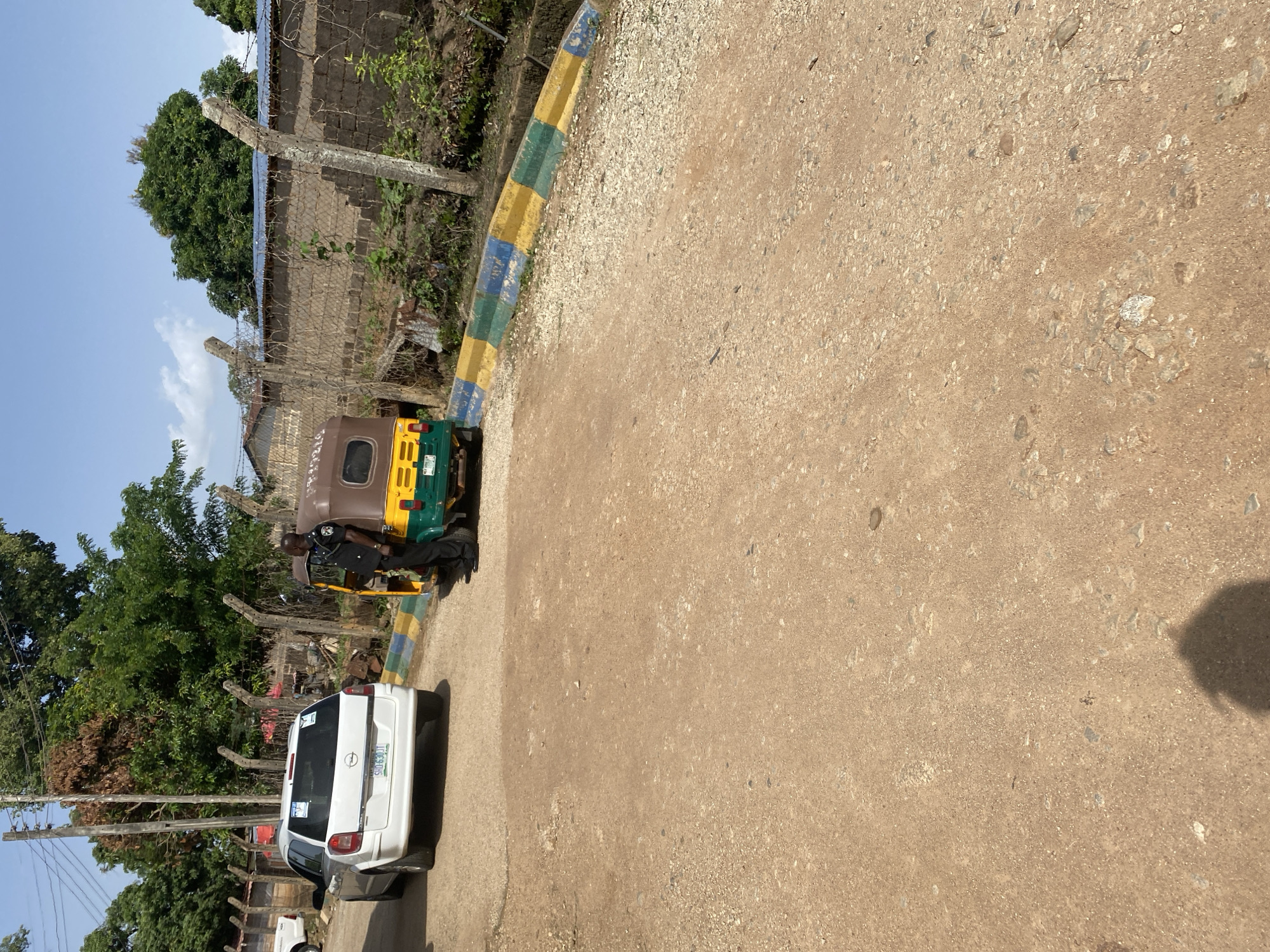 In Jos, Nigeria, a man in a dark uniform, likely a security officer, stands by a colorful auto-rickshaw. The auto-rickshaw is green and yellow, with brown detailing. Behind the officer and the auto-rickshaw, a white SUV is parked. The scene is set on a dirt road, bordered by a fence made of concrete posts and wire. Beyond the fence, a wall and lush green trees are visible under a clear blue sky. The ground is a mix of dirt and small stones.