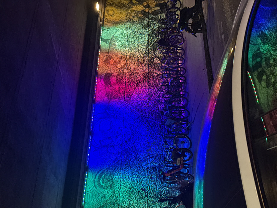 A vertical view displays a wall illuminated with a rainbow gradient. From top to bottom, the hues shift from yellow-green to pink, blue, and finally back to green. Silhouetted bicycles are aligned against the wall, casting dark outlines on the colorful backdrop. An indistinct image is reflected on a car window. The wall shows a faint decorative pattern. LED lights run along the edge.