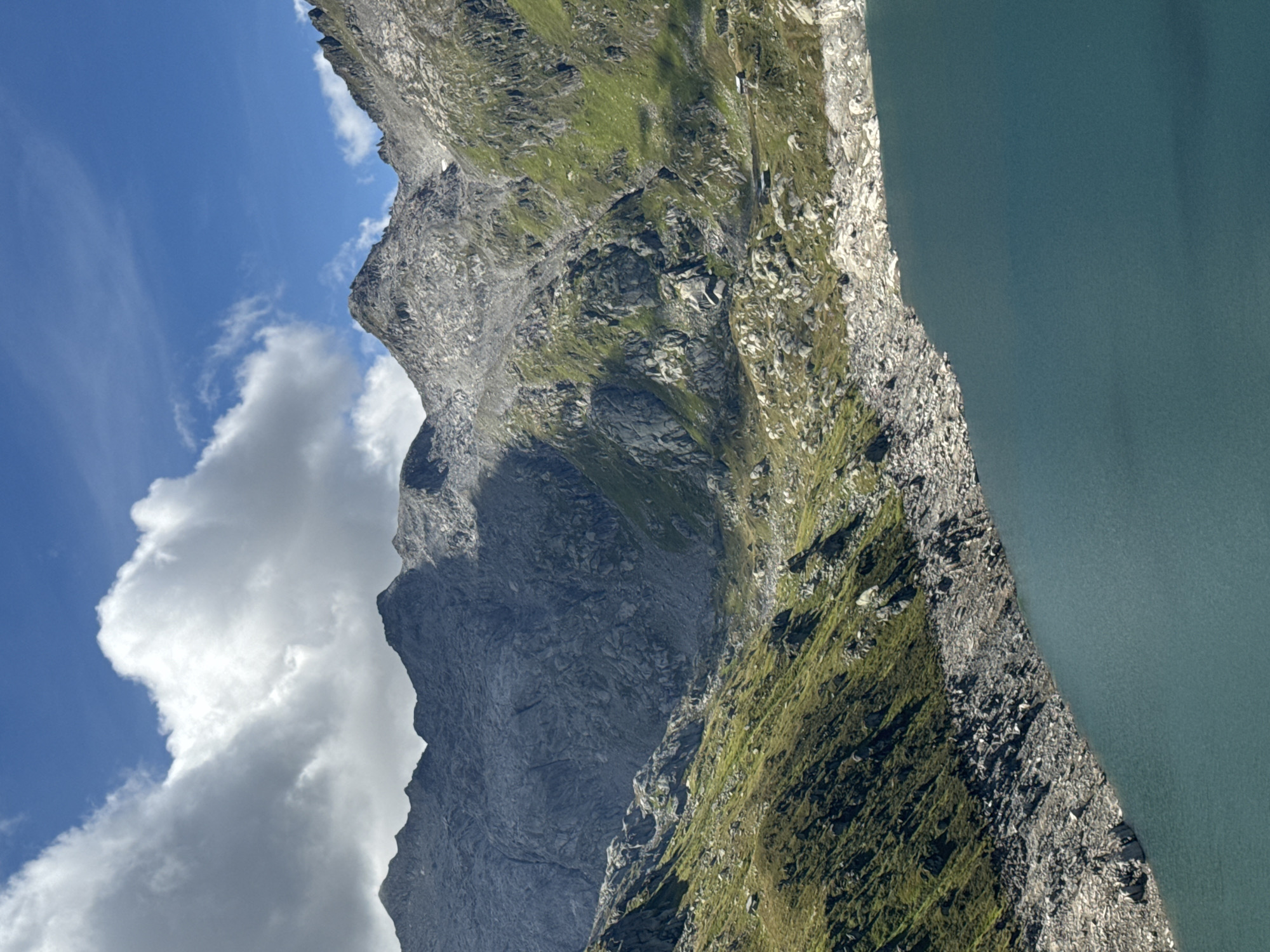 This is a landscape photograph of a lake and mountains. The lake is a deep blue-green color and fills the bottom right of the frame. The shore of the lake is lined with large, gray rocks and sparse green vegetation.
Behind the lake, the landscape rises steeply into rugged, gray mountains. The mountains are covered with patches of green grass and vegetation, especially on the lower slopes. There are significant rock formations and scree slopes visible on the mountainsides. The upper parts of the mountains are rocky and barren.
The sky is a bright blue with several large, white, fluffy clouds. Sunlight is casting shadows on the mountains, indicating a bright, sunny day. In the middle ground of the mountain slope, there appear to be a couple of very small, dark-colored structures, possibly huts or shelters.
The image is taken from a high vantage point, looking down and across the scene. The overall impression is one of a remote, natural alpine environment. The location is identified as a lake near Airolo, Switzerland.