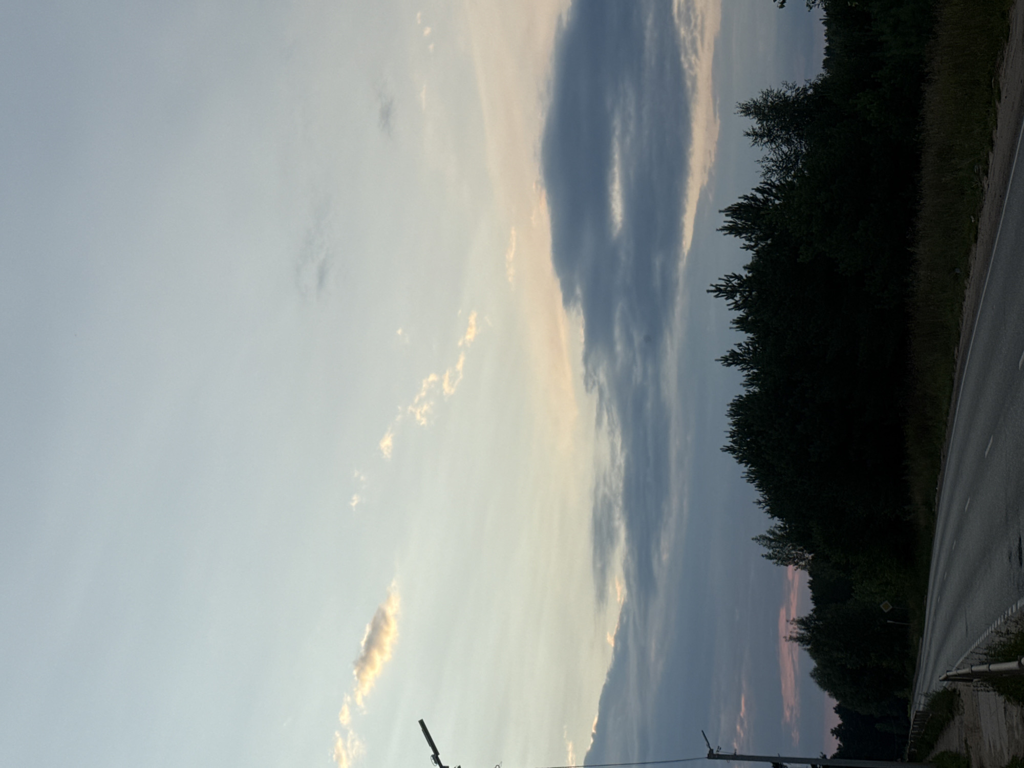 The image depicts a rural road in Chaikovsky, Russia, at dusk. The sky is a gradient of pale blue and dusky grey, with scattered clouds tinged with the soft hues of sunset. A line of dark, silhouetted pine trees forms a dense forest on the right side of the road. The road itself is paved and appears to be a two-lane highway, curving gently into the distance. A concrete sidewalk runs along the left side of the road, bordered by a metal guardrail. A solitary lamppost stands on the left side of the road, its dark silhouette stark against the sky. The overall atmosphere is tranquil and serene, with the fading light casting long shadows and creating a sense of quietude. There are no people or vehicles visible in the scene.
