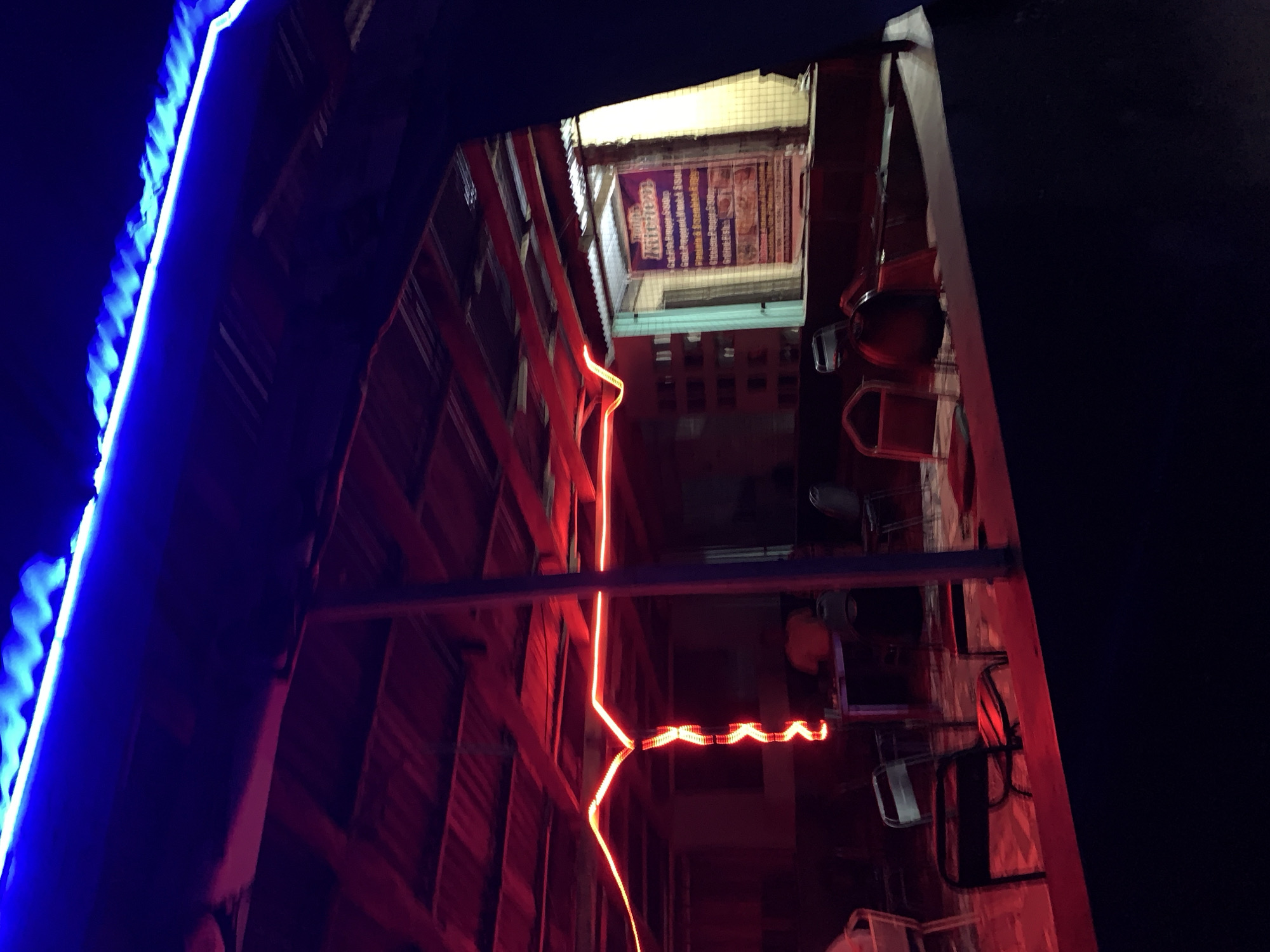 The image, shot from a low angle, depicts an alleyway at night, possibly in the city near New Stadium, Nigeria, based on the sign for "Kitchen" visible at the end of the alley. The alley is narrow, with wooden structures on either side. Blue LED lights adorn the upper left side of the alleyway. A red neon light runs along the right side of the wooden structure, leading to the end of the alley. The end of the alley appears to be lit from behind, revealing a sign that says "Kitchen." There are also some objects, potentially kitchenware or signage, hanging on the side.