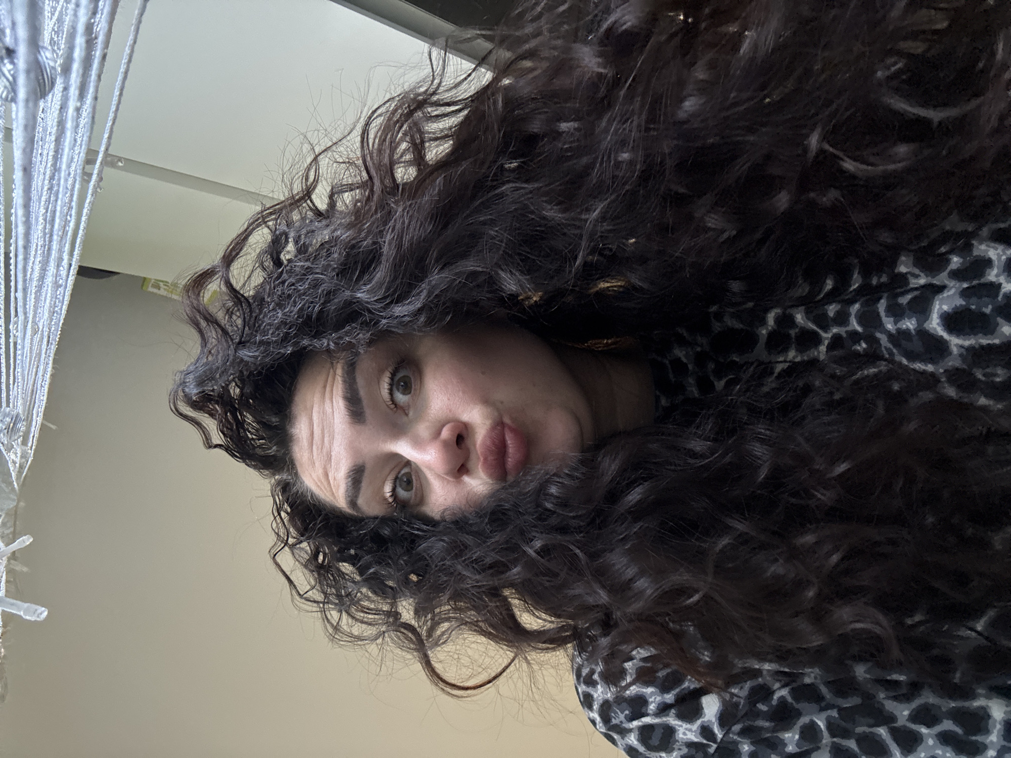 A woman with long, dark, curly hair is making a kissing face at the camera. She is wearing a black and gray leopard-print shirt. Her eyes are wide and her eyebrows are raised. She is in a room with beige walls and what appears to be a string of lights or a curtain hanging from the ceiling. The lighting is bright.