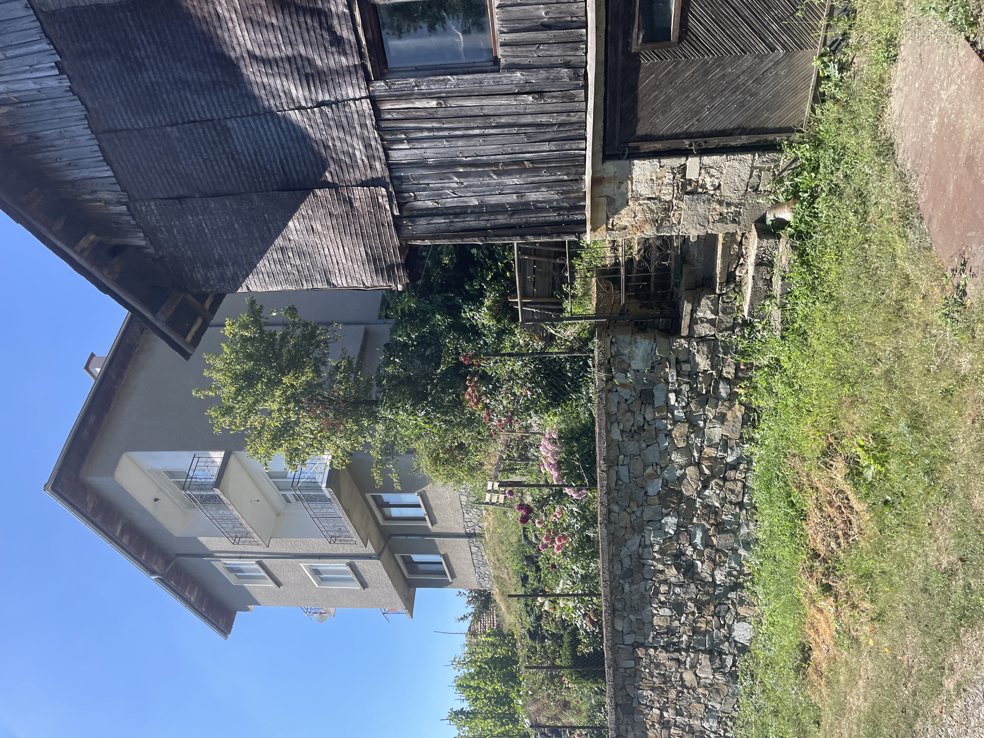 The image depicts a rustic scene in the village of Davidkovo, Bulgaria, on a clear, sunny day. Two adjacent buildings dominate the frame: a multi-story, grey, stucco building with balconies, and an older, weathered wooden structure with a corrugated roof. In the foreground, a stone wall runs horizontally, separating the grassy embankment from a vibrant flower garden featuring pink and purple blooms. A set of stone steps leads up from the lower level to the wooden building, which appears to be an outbuilding or garage with a large wooden door. The overall environment suggests a rural, slightly overgrown setting. There are no people visible in the image.