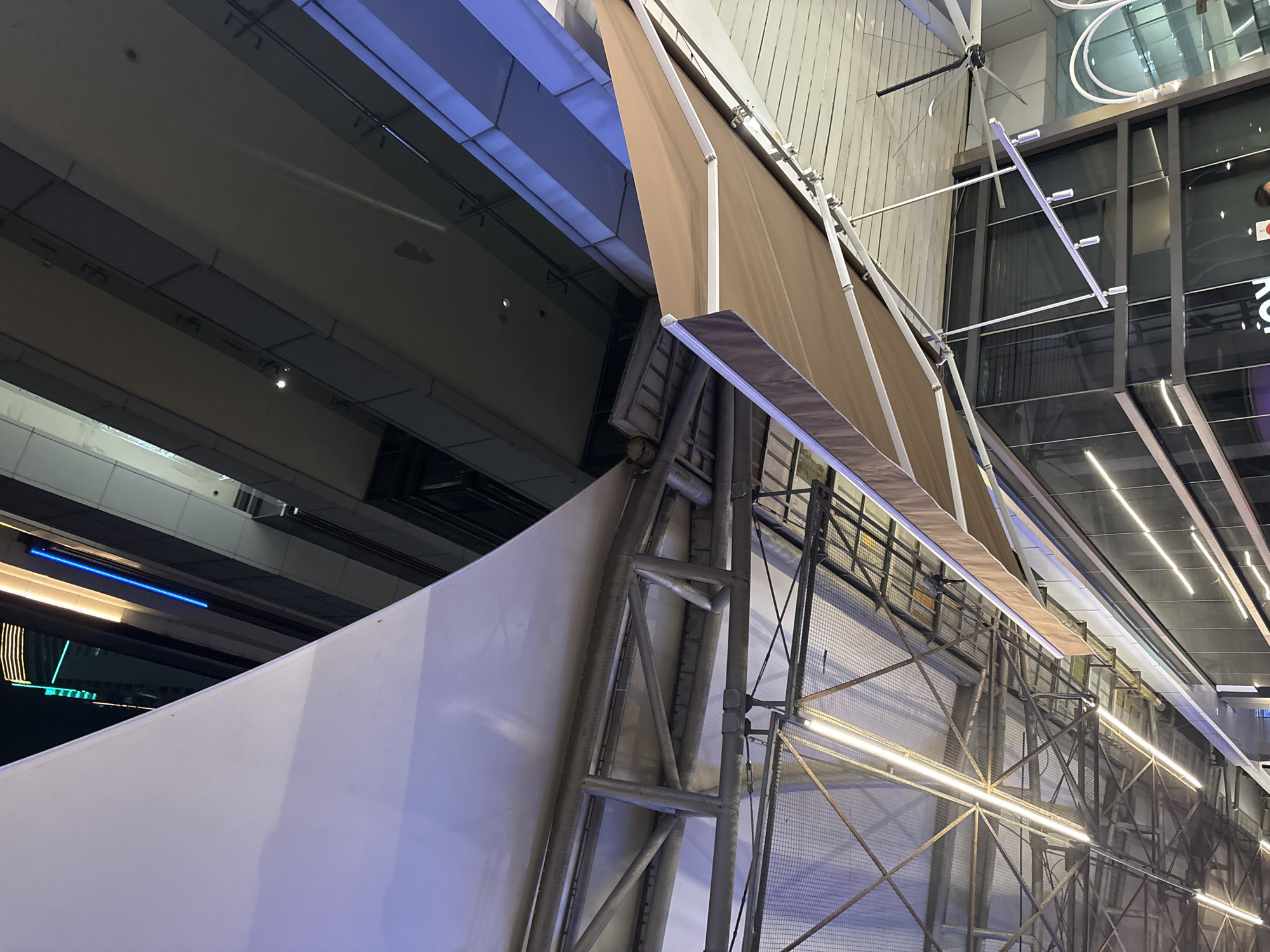 This is a low-angle, medium shot of the exterior of a modern building, likely in a city. The scene is illuminated by artificial lights, suggesting it is either evening or night. In the foreground, a large white fabric awning is partially visible, extending from the left side of the frame. Below it, a metal truss structure supports the awning and other architectural elements. A second, smaller, brown fabric awning is visible to the right, also supported by metal framework. The background features a multi-story building with a glass facade, reflecting the surrounding lights. Distinctive blue and yellow neon lights are visible on the facade of the background building, indicating a commercial or entertainment district. The overall impression is one of urban architecture and infrastructure. Without further information or context, pinpointing the exact location within North Jakarta, Indonesia is not possible based solely on the visual cues in the image.