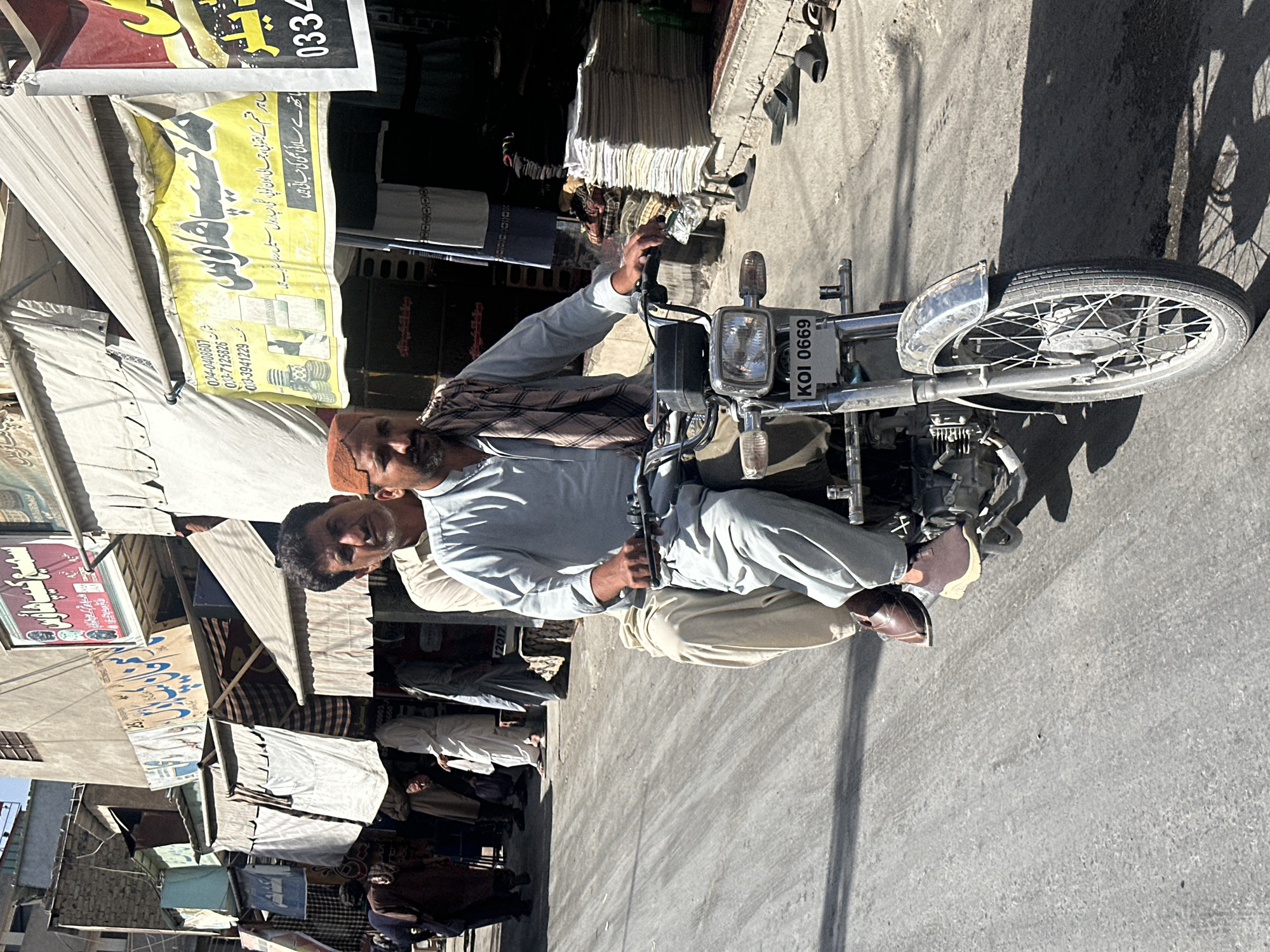 Two men are seated on a motorcycle with the license plate KOI 0669. The man in the front is wearing a light blue shirt, tan pants, and brown shoes. He is also wearing a scarf around his neck and a hat. The man in the back is wearing a light blue shirt and tan pants. The location appears to be Quetta City Tehsil, Pakistan, as indicated by the Urdu signage on the buildings. There are shops with awnings and merchandise displayed. Other pedestrians are visible in the background.