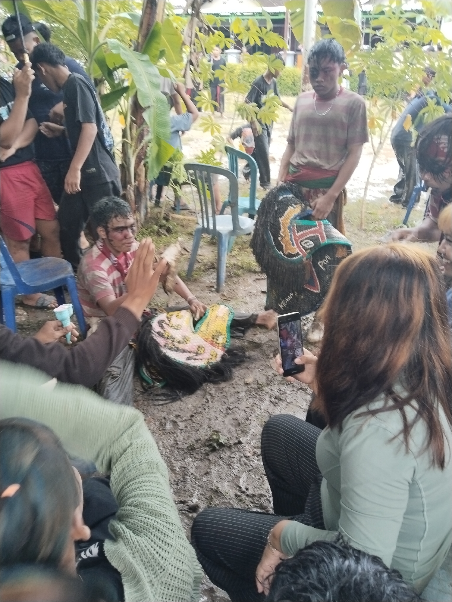 In a muddy outdoor area in Karang Tengah, Indonesia, a group of people gathers around two costumed figures. One figure, seated on the ground, wears a pink-striped shirt and smeared makeup, resting beside a large, colorful mask with black hair. Another person stands holding a similar, upright mask. A woman seated nearby holds a mobile phone, seemingly taking a picture of the scene. Additional individuals stand or sit around the perimeter, some observing, while others use umbrellas. Several blue plastic chairs are scattered across the muddy ground. Trees and vegetation surround the area, suggesting an outdoor gathering space.