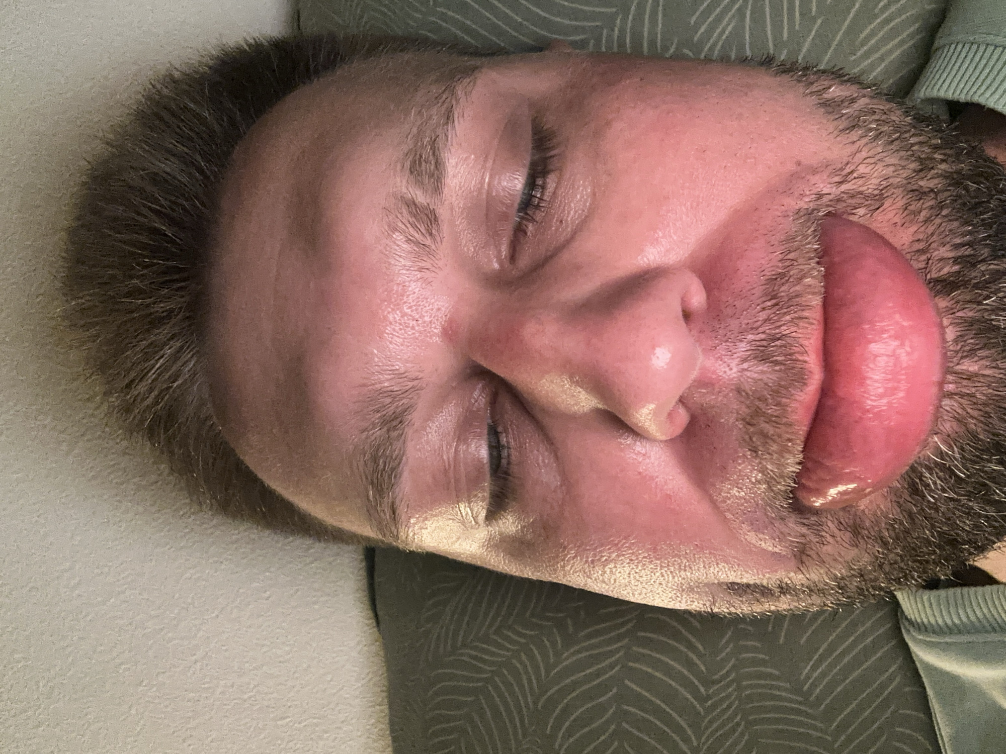 The image is a close-up, overhead shot of a person lying down with their eyes closed and tongue extended. The person has fair skin, short, brown hair, and stubble on their face. Their right eye is visible, with long eyelashes. The tongue is a prominent feature, appearing swollen and moist. The person is wearing a light green crew neck sweatshirt.
The background consists of two textured surfaces. To the left is a light-colored, textured surface, possibly a wall or fabric. To the right is a darker green surface with a repeating pattern of wavy lines.
The lighting appears to be from above, casting soft shadows and highlighting the glossy texture of the tongue and skin. The overall impression is one of relaxation or perhaps a playful pose. There are no visible textual elements in the image. The setting is likely indoors, given the lighting and soft background textures. The context provided places the image in Zvezdara Urban Municipality, Serbia.