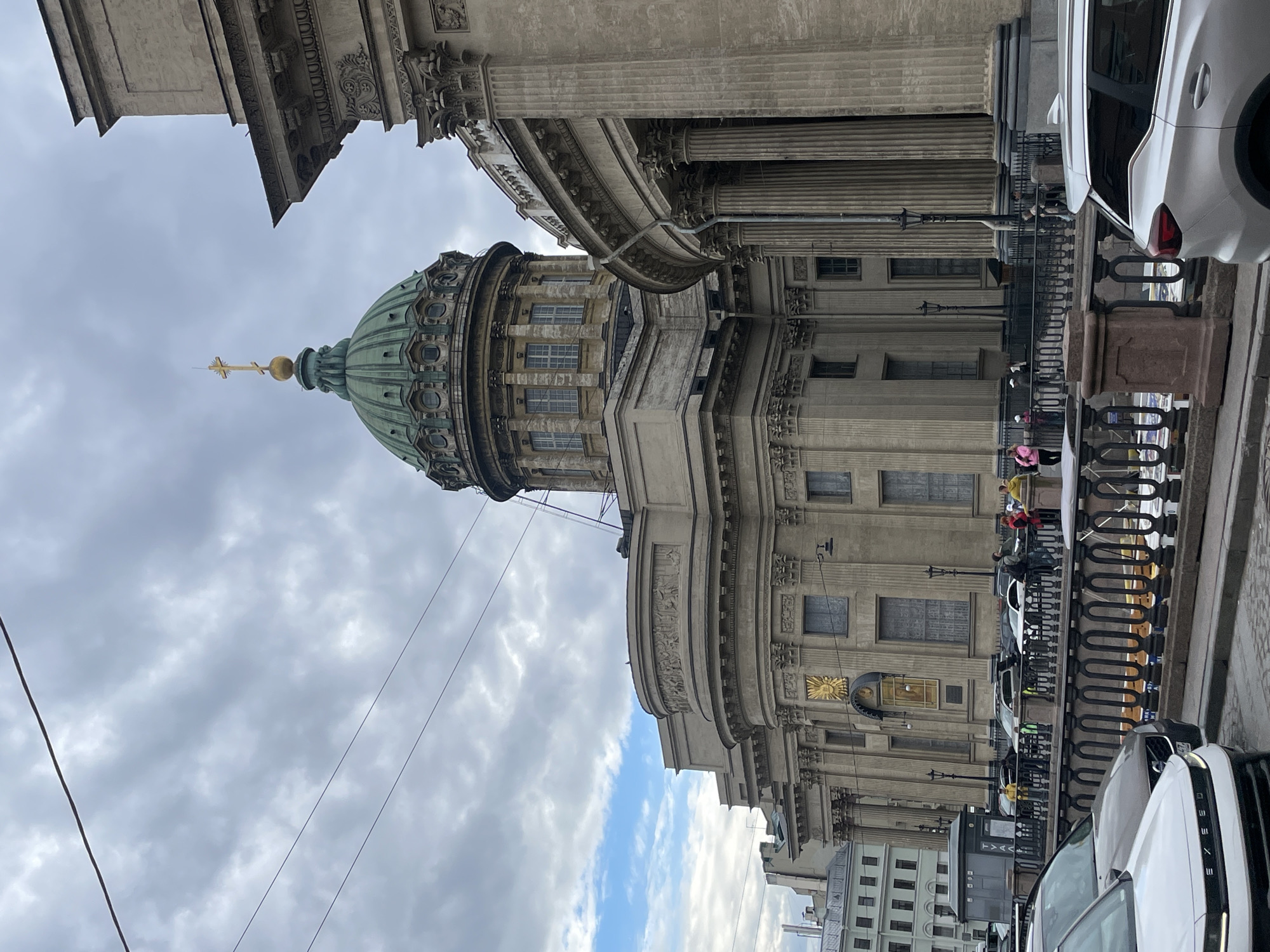The image shows a view of Kazan Cathedral in Saint Petersburg, Russia, on a cloudy day. The cathedral's prominent green dome with a golden cross is visible, as is its colonnade and neoclassical architecture. In the foreground, a street with cars and people is depicted. Several individuals are walking along the sidewalk and near the railing separating the street from a body of water. Cars, including a white SUV in the lower right corner and a white sedan in the lower left, are parked or moving on the street. The sky is overcast with patches of blue sky visible through the clouds. The overall scene captures a typical day in the historic city center.