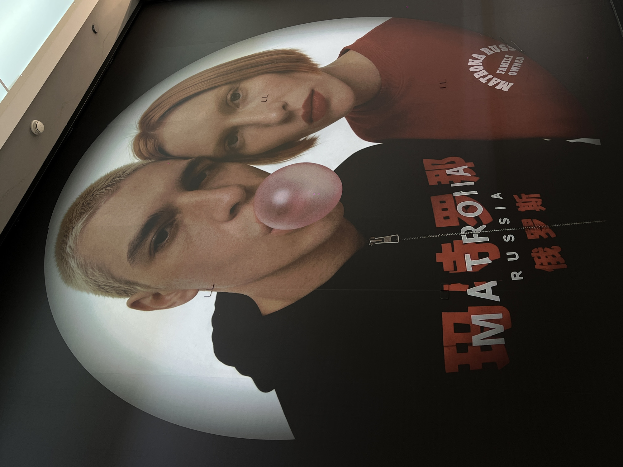 A large, stylized advertisement or poster on a wall features two individuals against a bright, circular white background, contrasted with a dark surrounding area. The image is tilted, with the subjects appearing diagonally.
The primary subject in the foreground is a person with short, light blonde hair and a distinct side part. They are shown from the chest up, wearing a black zippered hoodie. This individual is actively blowing a large, translucent pink bubblegum bubble. A small, dark cross earring is visible on their left ear.
Above and slightly behind this individual is another person with short, reddish-blonde hair and defined eyebrows. They are wearing a reddish-brown crewneck sweatshirt. This person has prominent red lipstick and a small, stylized "LL" visible on the bridge of their nose, possibly a tattoo or makeup.
Visible text on the clothing identifies brand names and origin. On the black hoodie, large red text reads "MATRONA RUSSIA" with the Cyrillic word "РУССИЯ" (Russia) also present. On the reddish-brown sweatshirt, text wrapped in an arc reads "MATRONA RUSSIA FAMILY OWNED" in white. Small "LL" symbols are also present on the red sweatshirt.
The setting appears to be indoors, possibly a commercial space, with architectural elements and what looks like a fire alarm or smoke detector visible in the top left corner of the original image, hinting at a modern building. The overall mood is cool and contemporary, characteristic of fashion advertising.