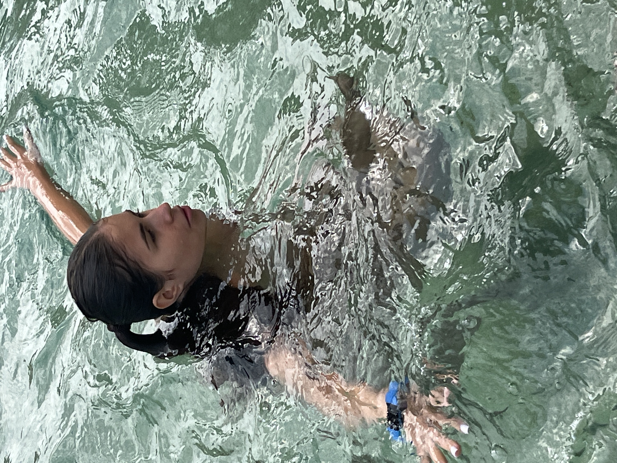 A young woman with dark hair in a ponytail is in the water. She is wearing a dark swimsuit and a blue and black band on her wrist. Her right arm is extended upwards and her left arm is bent, with her hand near her chest. She is looking to the right with her mouth slightly open. The water is clear and green, with ripples and reflections on the surface. The background is out of focus. There are no identifiable landmarks or features in the image that would indicate a specific location within Poltava, Ukraine.