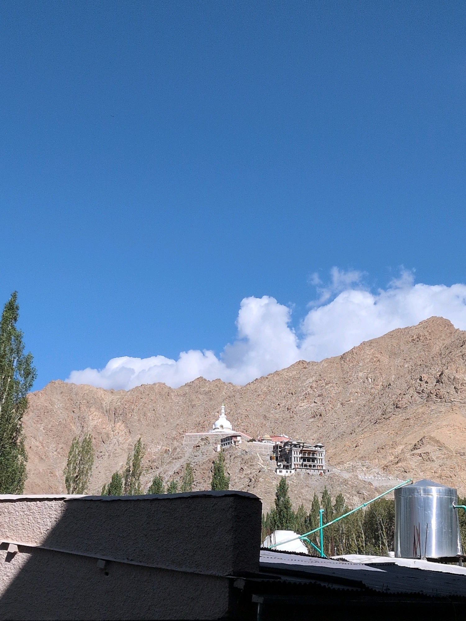 The image captures a panoramic view of a mountainous landscape in Leh, India, dominated by a clear blue sky with scattered white clouds. In the foreground, a textured, light-colored wall casts a shadow, suggesting it is part of a building's exterior. Beyond this, a corrugated roof is visible, along with a large, cylindrical silver water tank.
The mid-ground reveals a range of arid, brown mountains. Nestled on one of the slopes is a prominent white stupa, a Buddhist monument, with a multi-story building complex built behind and to the side of it. A winding road leads up to these structures. A line of slender, green trees stretches across the base of the mountains, varying in height and density.
The background is defined by the imposing, rocky mountains, their surfaces textured and devoid of significant vegetation, characteristic of the high-altitude desert environment of Leh. The overall scene depicts a serene and majestic landscape, highlighting the unique architecture and natural beauty of the region.