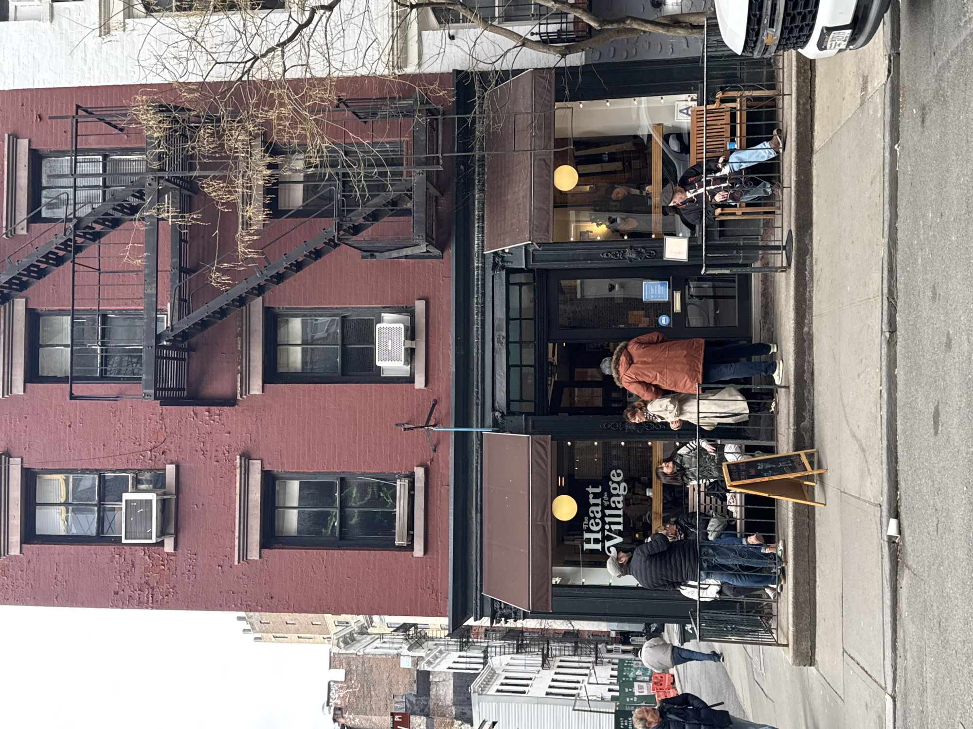 This image depicts a street scene in Greenwich Village, New York City, United States of America. The central subject is a three-story red brick building with visible paint deterioration on its facade. The ground floor houses a commercial establishment named "The Heart of the Village," identifiable by its sign featuring a circular yellow light emblem, situated beneath a dark brown awning.
The building exhibits dark-framed windows on all floors; two window-mounted air conditioning units are installed on the second and third stories. A black metal fire escape is affixed to the right side of the brick facade, extending upwards from the second level. To the immediate right, a portion of an adjacent white brick building is visible, and a bare tree with sparse branches partially obscures the fire escape and building.
On the sidewalk in front of "The Heart of the Village," a black metal railing encloses an outdoor seating area. Within this area, a minimum of eight individuals are present. Several are standing and interacting, including one male in an orange hooded jacket and blue jeans, facing a female in a light-colored coat and scarf. Another male in a grey cap and dark puffer jacket stands to the left of this interaction. Other individuals are seated on wooden benches positioned behind the railing. An A-frame chalkboard sign is placed on the sidewalk near the railing.
A white paper cup is discarded on the asphalt curb. To the left, a male subject in a dark jacket is walking along the sidewalk. To the right, the front portion of a white Chevrolet pickup truck is parked at the curb. Through the storefront windows, additional individuals are visible inside the establishment. The background to the left reveals more multi-story urban buildings with fire escapes under an overcast sky.