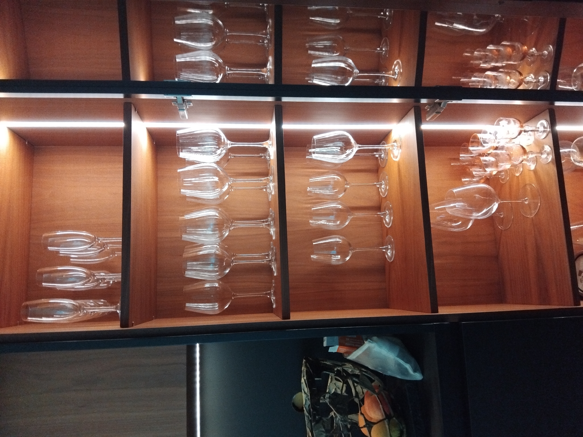 The image shows a wooden cabinet with several shelves displaying glassware. On the top shelf, there are tall, slender champagne flutes. The middle shelf has a collection of wine glasses with wider bowls. The bottom shelf holds a variety of smaller glasses, possibly for liqueurs or digestifs. The cabinet is illuminated by vertical strip lights, creating a warm glow. In the lower left corner, part of a dark countertop is visible, with a woven basket containing fruit. The cabinet appears to be located in a domestic setting, likely a kitchen or dining area. The city of Vila Nova de Famalicão, Portugal, is mentioned in the prompt, but there is no visual information in the image to confirm this specific location.