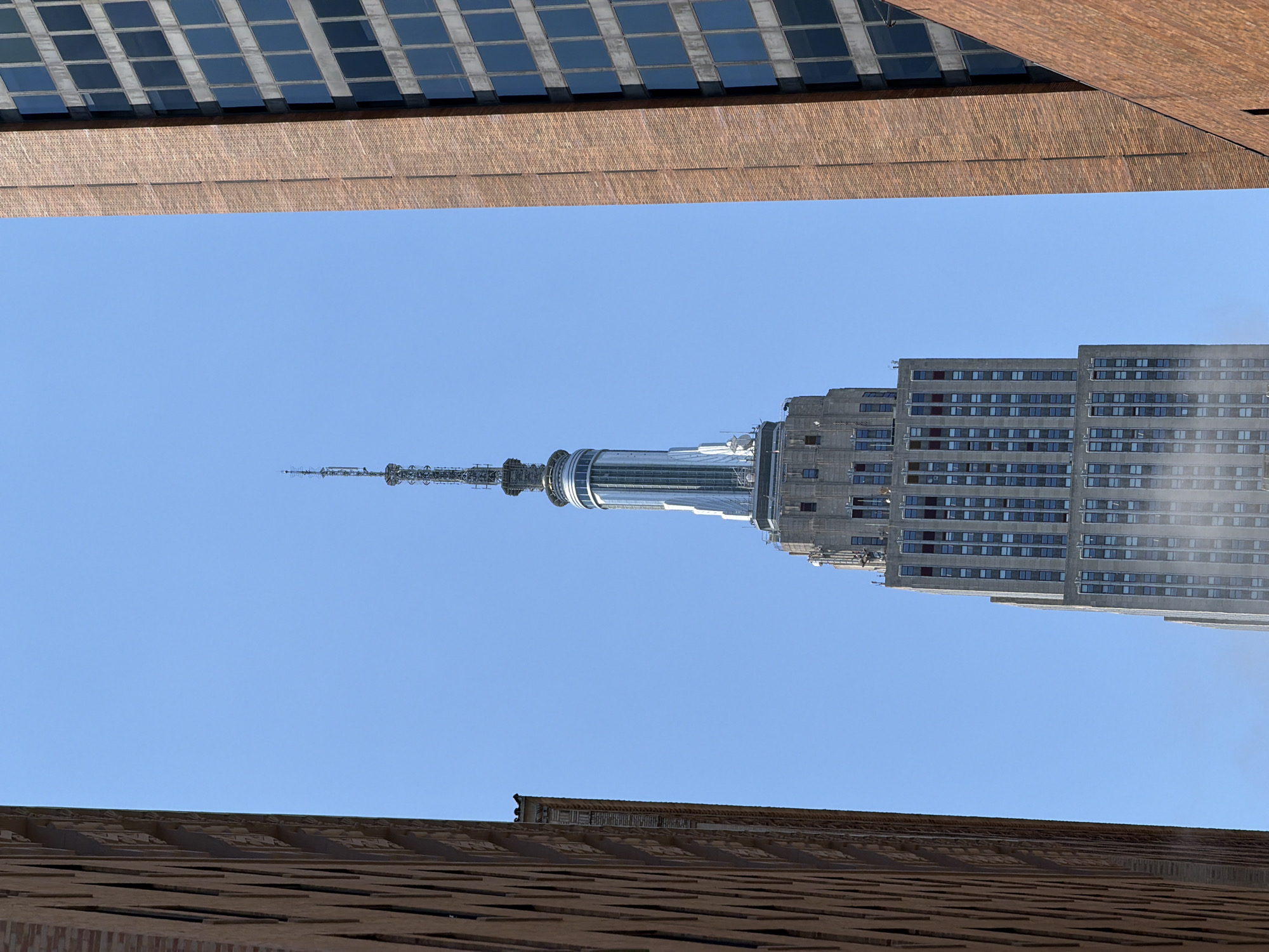 The image captures a striking view of the Empire State Building, a prominent skyscraper in New York City, USA. The photograph is oriented vertically, with the top of the image showing the upper portion of two other buildings, one with a facade of brown brick and the other with a grid of dark windows. Below this, the sky is a clear, bright blue, indicating a sunny day.
The Empire State Building dominates the central part of the frame, its distinctive spire reaching towards the sky. The building's iconic art deco architecture is visible, with its tiered structure and numerous windows. Construction or maintenance equipment can be seen near the spire, suggesting ongoing work on the landmark.
At the bottom of the image, another building with a textured brown facade, possibly brick or a similar material, is visible. This building is angled and appears to be a lower structure in the foreground.
The overall impression is one of an urban landscape on a clear day, highlighting the architectural grandeur of the Empire State Building within the context of the city. The image provides a unique perspective, looking up at the skyscraper from a lower vantage point and framed by other buildings. There is no visible text in the image.