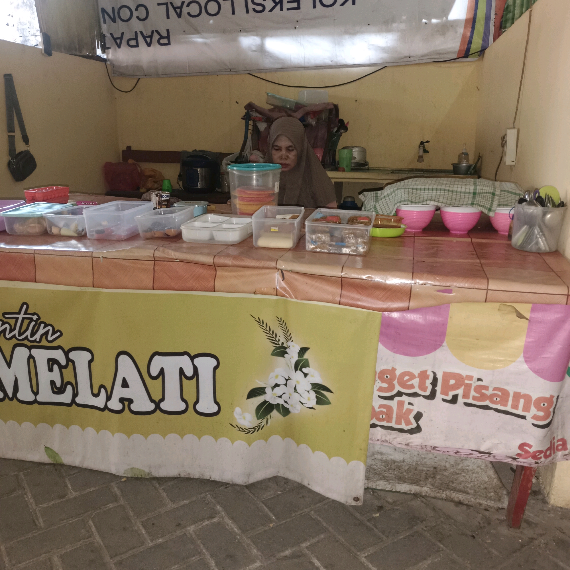 The image depicts a street food stall in Surabaya, Indonesia. A woman wearing a brown hijab stands behind a table laden with food containers. The stall features a yellow banner with white lettering that reads "MELATI", suggesting it is a stall selling jasmine-related products or services. Another banner with the words "get Pisang" and "Sedia" is visible to the right, indicating banana snacks are available. In the background, a banner with Indonesian text, possibly related to local events or government announcements, hangs above the stall. The overall scene is a typical bustling street market environment.