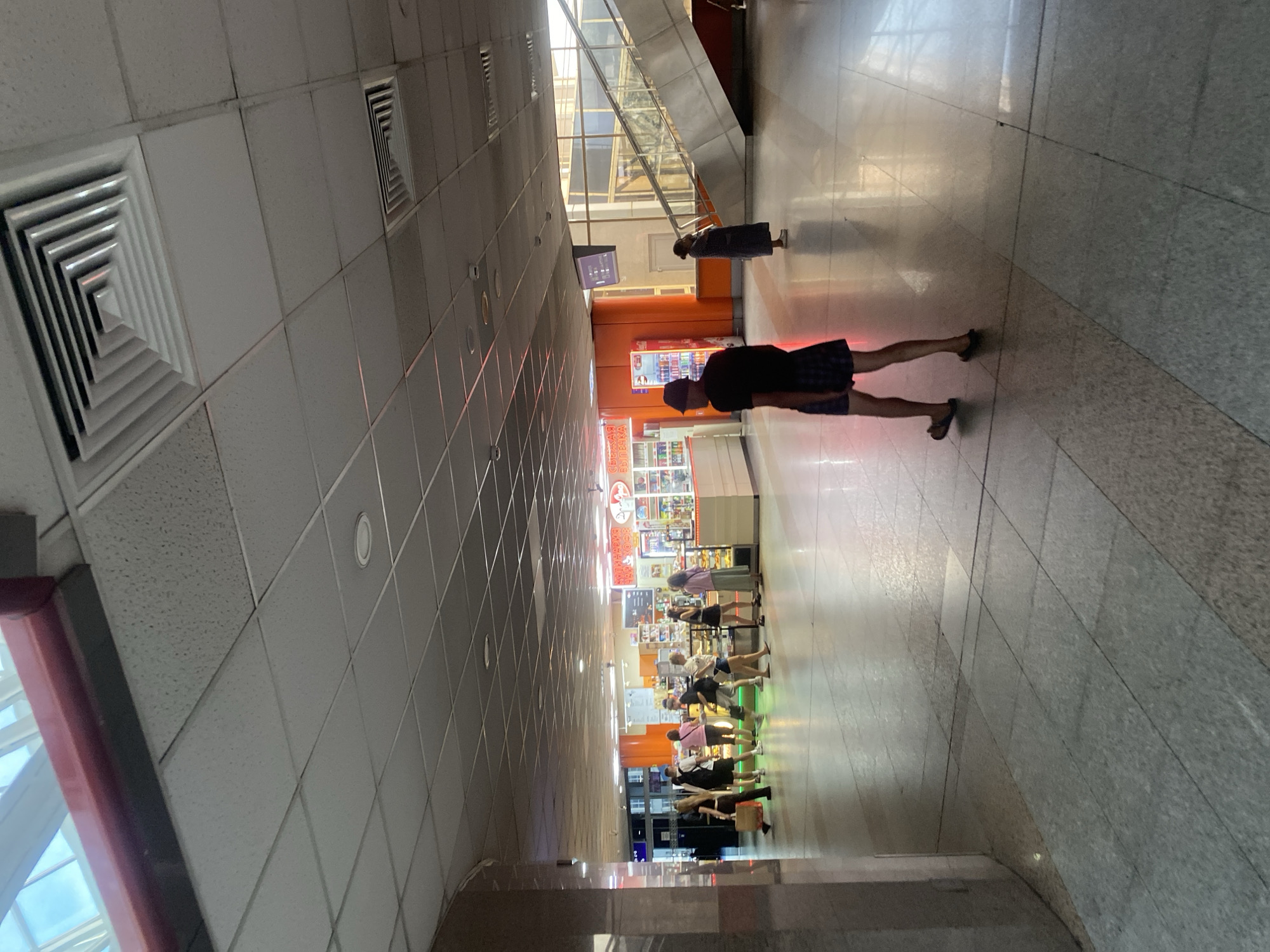 The image depicts the interior of a public space, possibly a transportation hub or shopping mall, in Minsk, Belarus. In the foreground, a person wearing a dark t-shirt and plaid shorts walks away from the camera. Further into the scene, a group of individuals is gathered around what appears to be a food kiosk or convenience store. To the right, an escalator is visible, with a person standing near its base. The environment features a tiled floor, a drop ceiling with ventilation grates, and bright lighting. The overall impression is of a busy, functional indoor area.