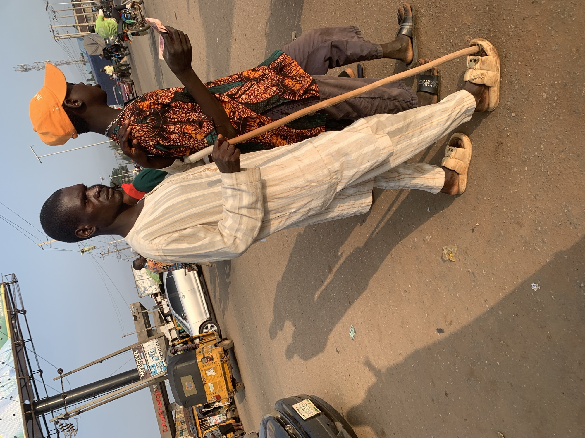 A man wearing a white and cream striped robe and sandals, leaning on a wooden stick, stands in the foreground. To his right, another man in a colorful patterned shirt and orange cap stands with his arm on the first man's shoulder, holding a few bills in his hand. They are on a dirt road in Shagamu, Nigeria. In the background, a yellow auto-rickshaw is visible, along with other vehicles, buildings, and a large billboard that reads "TotalEnergies." The scene is illuminated by the warm light of what appears to be late afternoon or early morning.