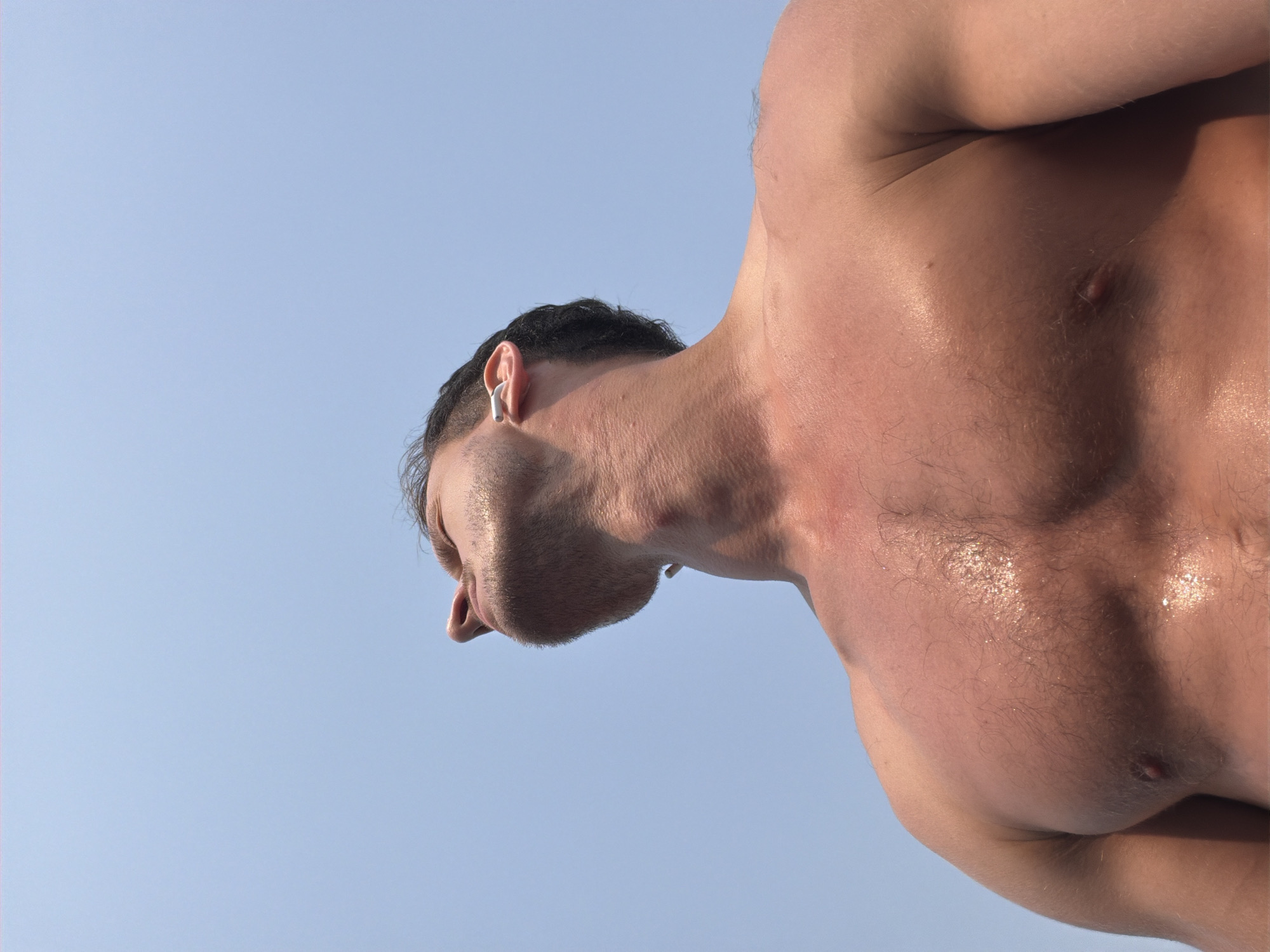 The image depicts a close-up, low-angle shot of a man's upper body and head against a pale blue sky. He is shirtless, showing his chest, neck, and part of his face from below. The man has short, dark hair and is wearing a white wireless earbud in his left ear. His skin appears slightly tanned and glistening. The location is not discernible from the image; therefore, it is impossible to determine if the image was taken in Đà Nẵng, Vietnam.
