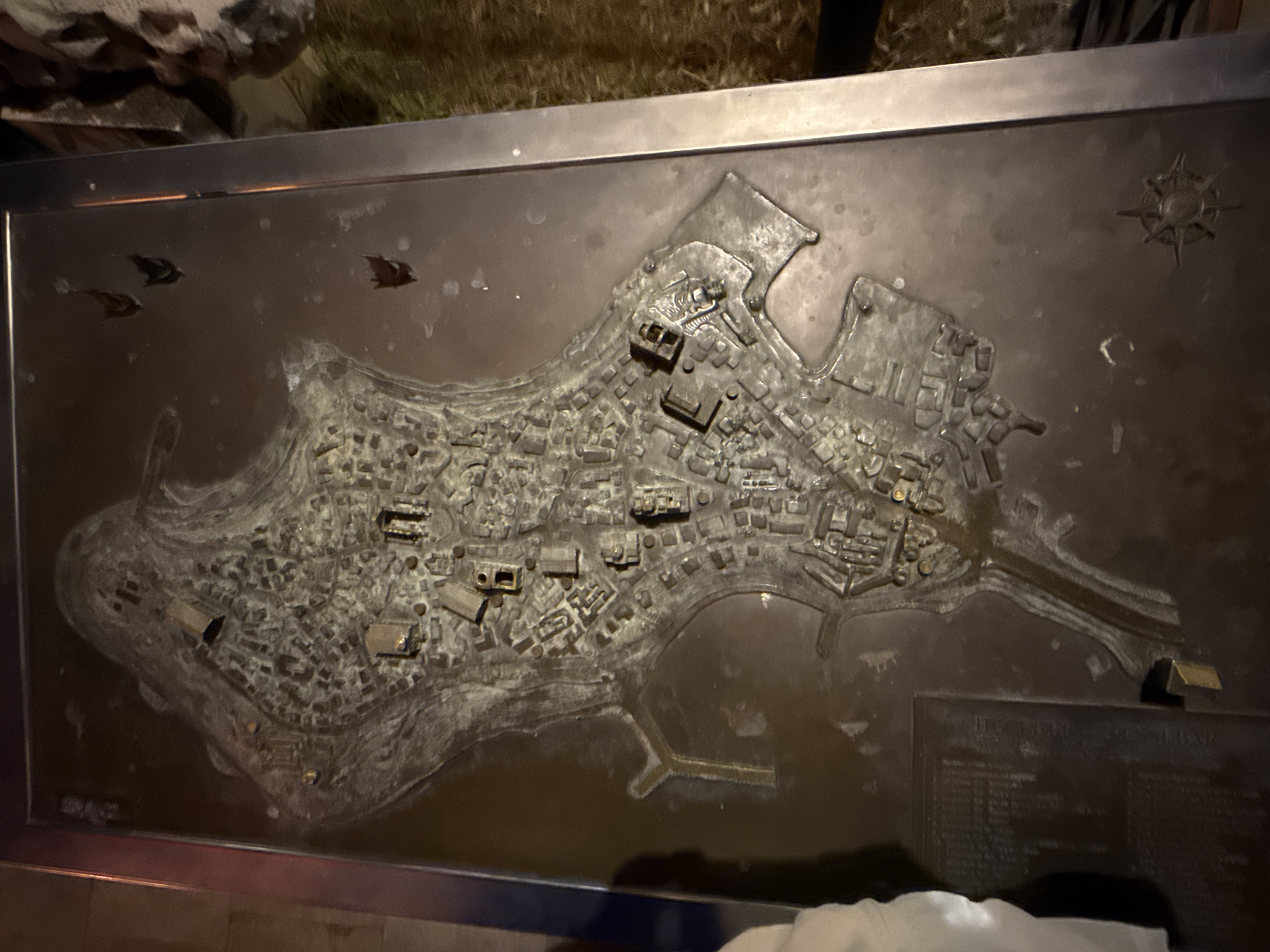 The image shows a detailed bronze relief map of Nesebar, Bulgaria. The map depicts the Old Town peninsula with its buildings, streets, and harbors. Several prominent structures, such as churches and fortresses, are recognizable in relief. The map is mounted on a dark metal base with a metallic frame. In the bottom left corner, a portion of a plaque with text is visible, suggesting it provides information about the map or the city. The surrounding environment appears to be outdoors, with some vegetation and a stone surface visible in the background. The lighting suggests it might be evening or nighttime, with artificial light illuminating the map.