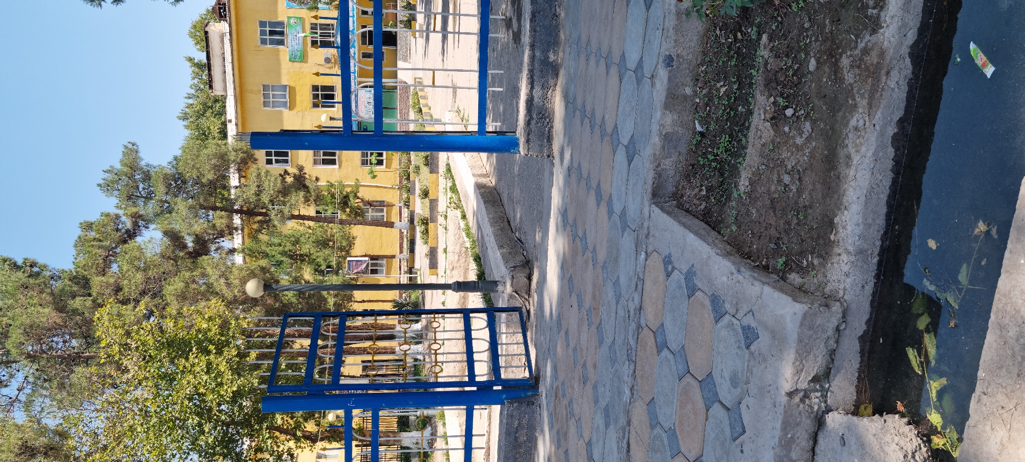 The image shows an exterior scene on a sunny day in the city of Ismoili Somoni, Tajikistan. In the foreground, a water channel is visible with some debris floating in it. The channel is bordered by concrete. Adjacent to the water channel is a paved pathway composed of hexagonal and diamond-shaped concrete tiles in beige and grey.
The pathway leads to an open gateway with blue metal bars. The gateway is flanked by a lamppost with a spherical light fixture. Behind the gate, a white metal fence is visible, and beyond that, several trees with lush green foliage.
In the background, a bright yellow building stands with multiple windows. Some windows have green banners or posters on them. The building appears to be a school or public institution. Trees are also planted in front of the building, with their trunks painted white at the base. The sky above is clear and blue. The overall scene depicts a public outdoor space with a building and surrounding greenery.