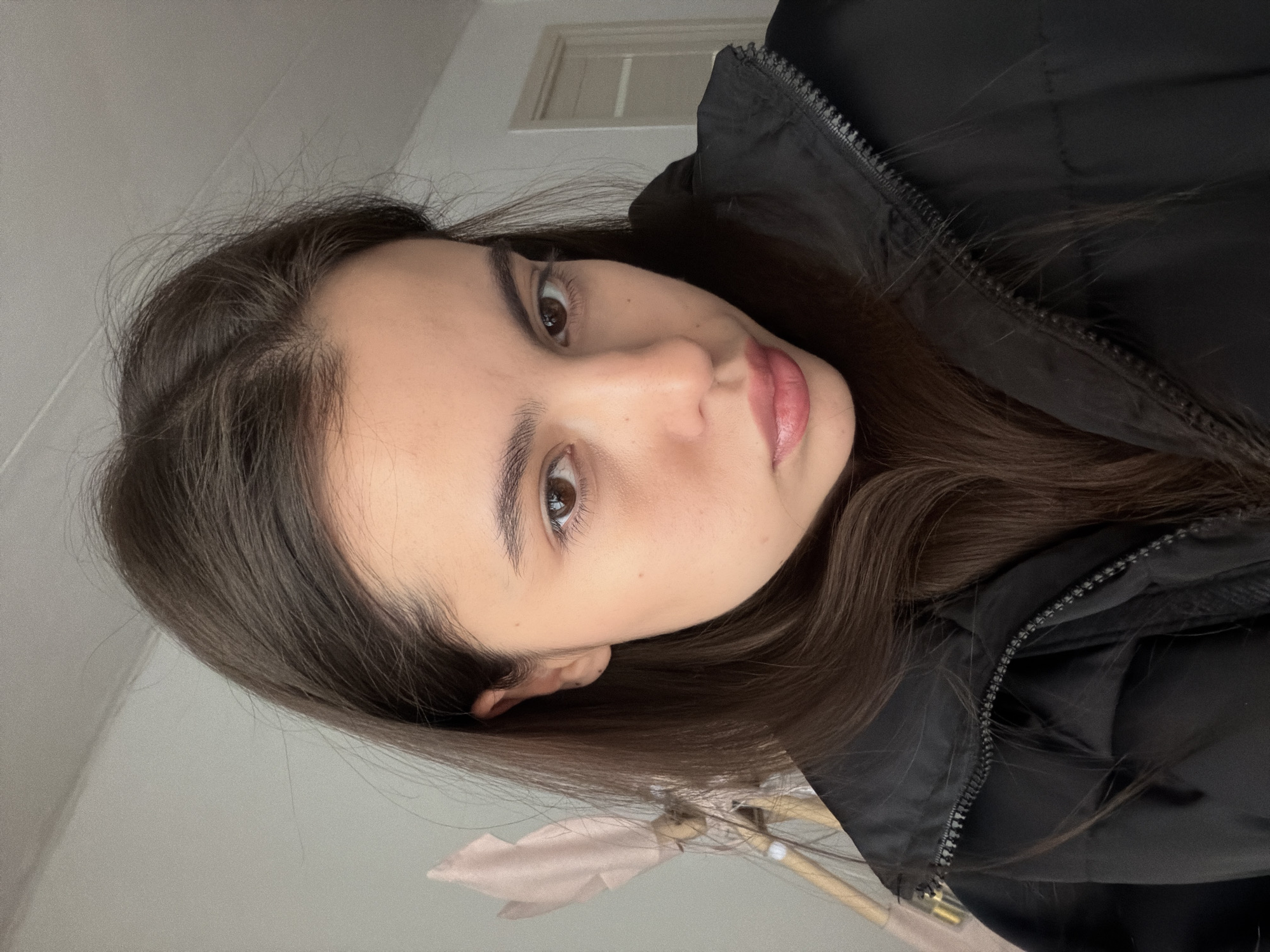 A medium shot shows a young woman in a dark jacket. Her hair is dark brown and her eyes are brown. She is looking at the camera. The background is a light gray wall and a window. The photo was likely taken indoors. The city of Chaikovsky, Russia is not visible.