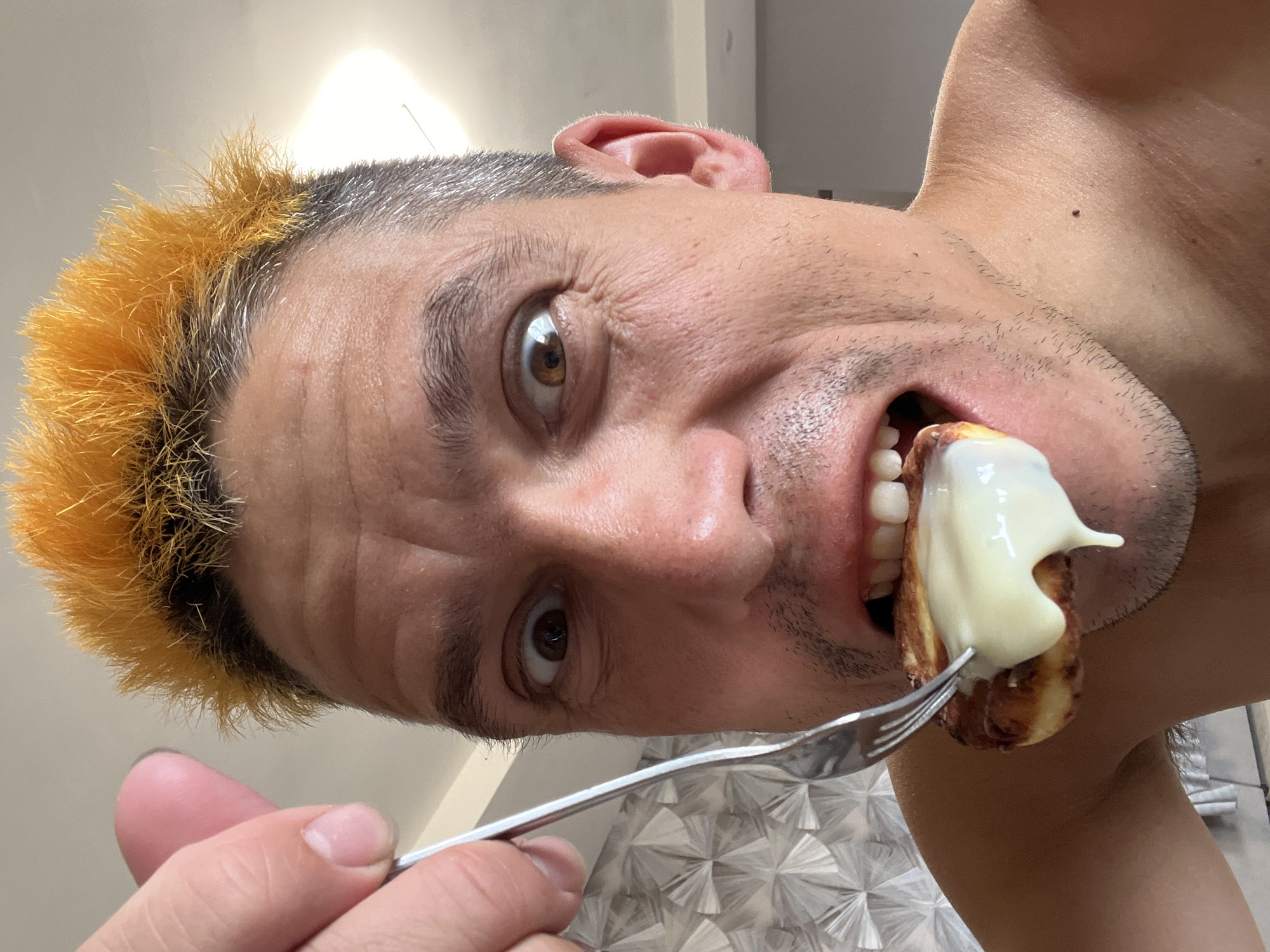 A close-up, low-angle, portrait orientation selfie shows a man with orange-tipped hair in Chaikovsky, Russia. He is about to eat a piece of food, possibly a pastry, that is covered in a white sauce and is held by a fork. The man's eyes are wide, and he has a playful or surprised expression. He has stubble on his chin and neck. The background is indistinct, but appears to be an indoor setting with a light-colored wall and a patterned surface that may be a tablecloth or placemat. The lighting suggests it is daytime, possibly indoors with artificial light. The image is slightly blurry, and the man's head is tilted to the side.
