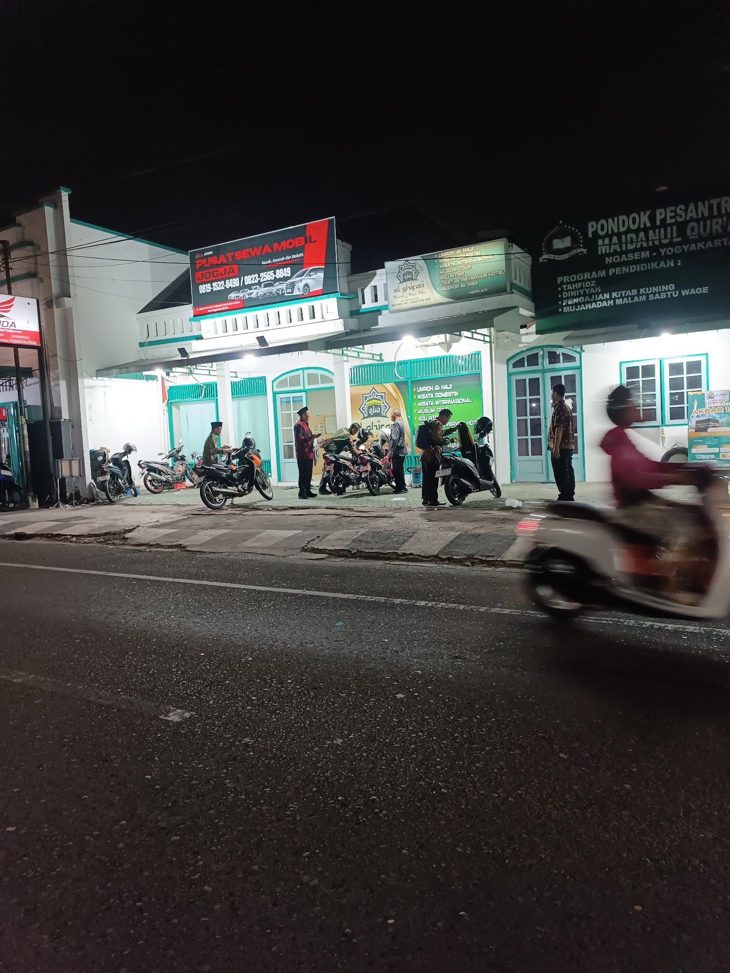 The image captures a nighttime scene on a street in Yogyakarta, Indonesia, likely in an area with commercial establishments. The foreground shows a blurred scooter in motion, indicating the photo was taken from a moving vehicle or with a slower shutter speed. The midground features a row of parked motorcycles and a building with several signs.
One prominent sign on the building advertises "PUSAT SEWA MOBIL JOGJA," a car rental service, with contact information and images of cars. Another sign above it is red and black, also related to car rental. To the right, a larger dark green sign displays "PONDOK PESANTREN MAIDANUL QUR'AN NGASAM - YOGYAKARTA" and lists "PROGRAM PENDIDIKAN" with items like "TAHFIDZ," "DINIYYAH," "PENGAJIAN KITAB KUNING," and "MUJAHADAH MALAM SABTU WAGE." Next to this, a lighter green sign with Arabic script and "al ghifari" text lists services such as "UMROH & HAJI," "WISATA DOMESTIK," "WISATA INTERNASIONAL," "ALBUM TOUR," "EDUKINTA," "TIKET & RESERVASI HOTEL," "PASPORT & VISA."
Several men are visible in the midground, some standing near the motorcycles and the building entrance, appearing to be interacting or waiting. The building itself has a light-colored facade with teal-colored window frames and arches, suggesting it might be a mosque or an educational institution. The street is paved and shows pedestrian crossings. The overall impression is a busy urban street at night, with businesses and a religious educational institution operating side-by-side.