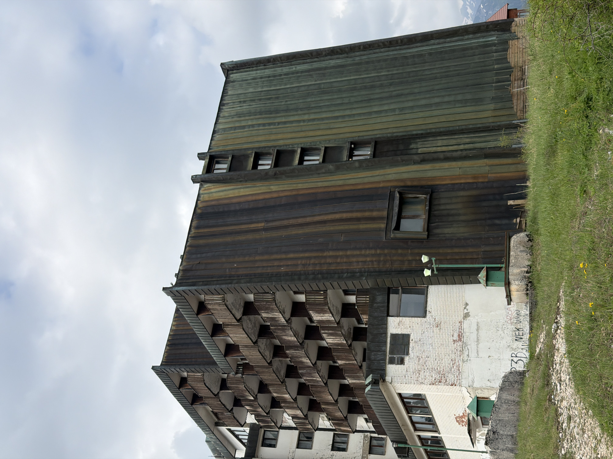 An exterior view of the partially dilapidated Hotel Durmitor in Žabljak, Montenegro. The hotel displays a Brutalist architectural style with horizontal wood siding on the upper floors and sawtooth balconies. The lower portion of the building is constructed of weathered white brick with some graffiti present. Green grass and a dirt path are visible to the right. A gray sky is above.