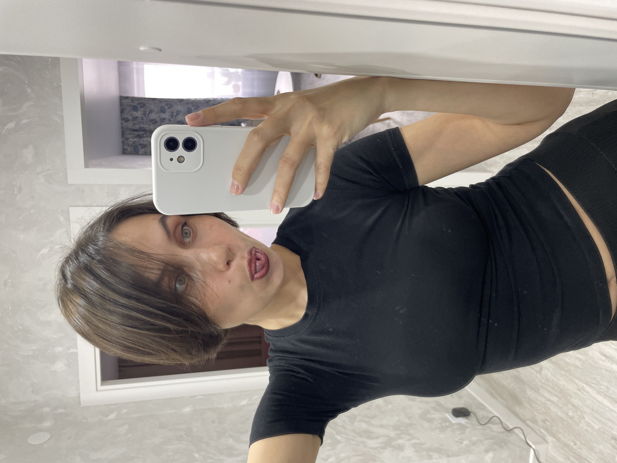 A woman with short brown hair and green eyes is taking a selfie in what appears to be a bathroom. She is wearing a black t-shirt and black bottoms. She is sticking her tongue out. The background includes a marble-patterned wall and a doorway or window with curtains. There is no information provided to determine the location as Izhevsk, Russia.