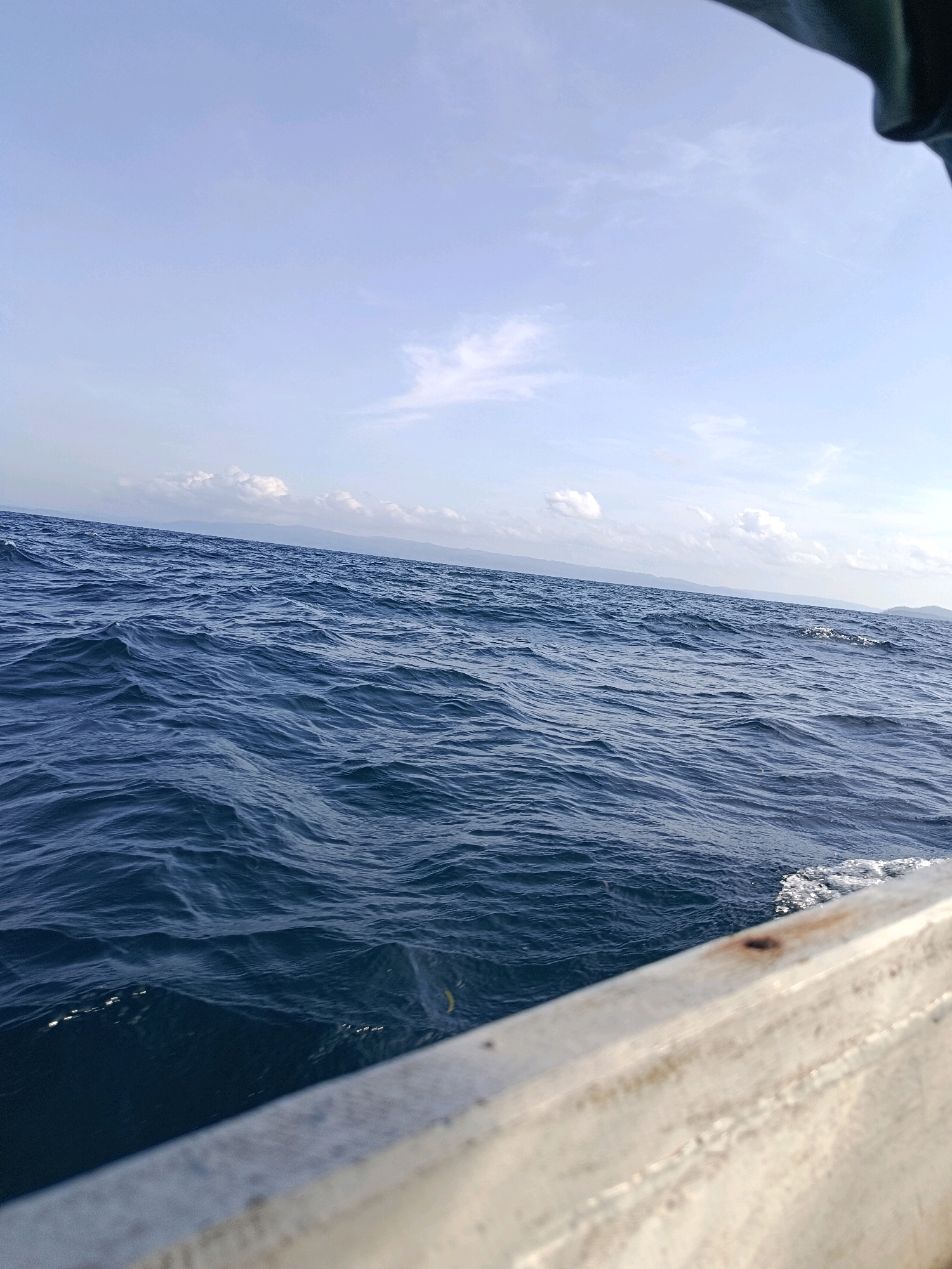 A wide-angle, low-angle shot captures the choppy, deep blue ocean from the perspective of a boat in Lompio, Indonesia. The water occupies the majority of the frame, with gentle waves creating a textured surface that reflects the bright, pale blue sky. Wispy white clouds are scattered across the sky, and a faint line of distant land, possibly mountains or hills, can be seen on the horizon. The edge of a metallic boat railing, weathered and a light grey color, occupies the bottom right of the image, extending diagonally into the frame. The scene is peaceful and natural, conveying a sense of being at sea under a clear sky.