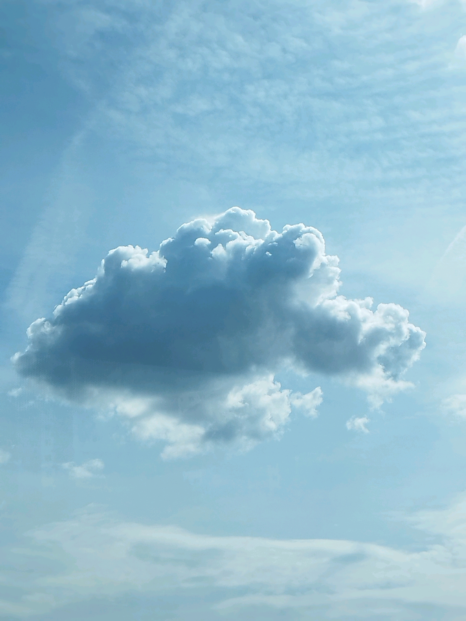 The image shows a sky with a large, dense cloud in the center, illuminated by sunlight. The sky is a pale blue, with wispy clouds and contrails visible in the background. The main cloud is white and gray, with shadowed areas indicating its depth and form. There are no people, structures, or any other objects visible in the image, nor any indication of a specific location within the city of Bragadiru, Romania. The scene is purely an atmospheric view of the sky.