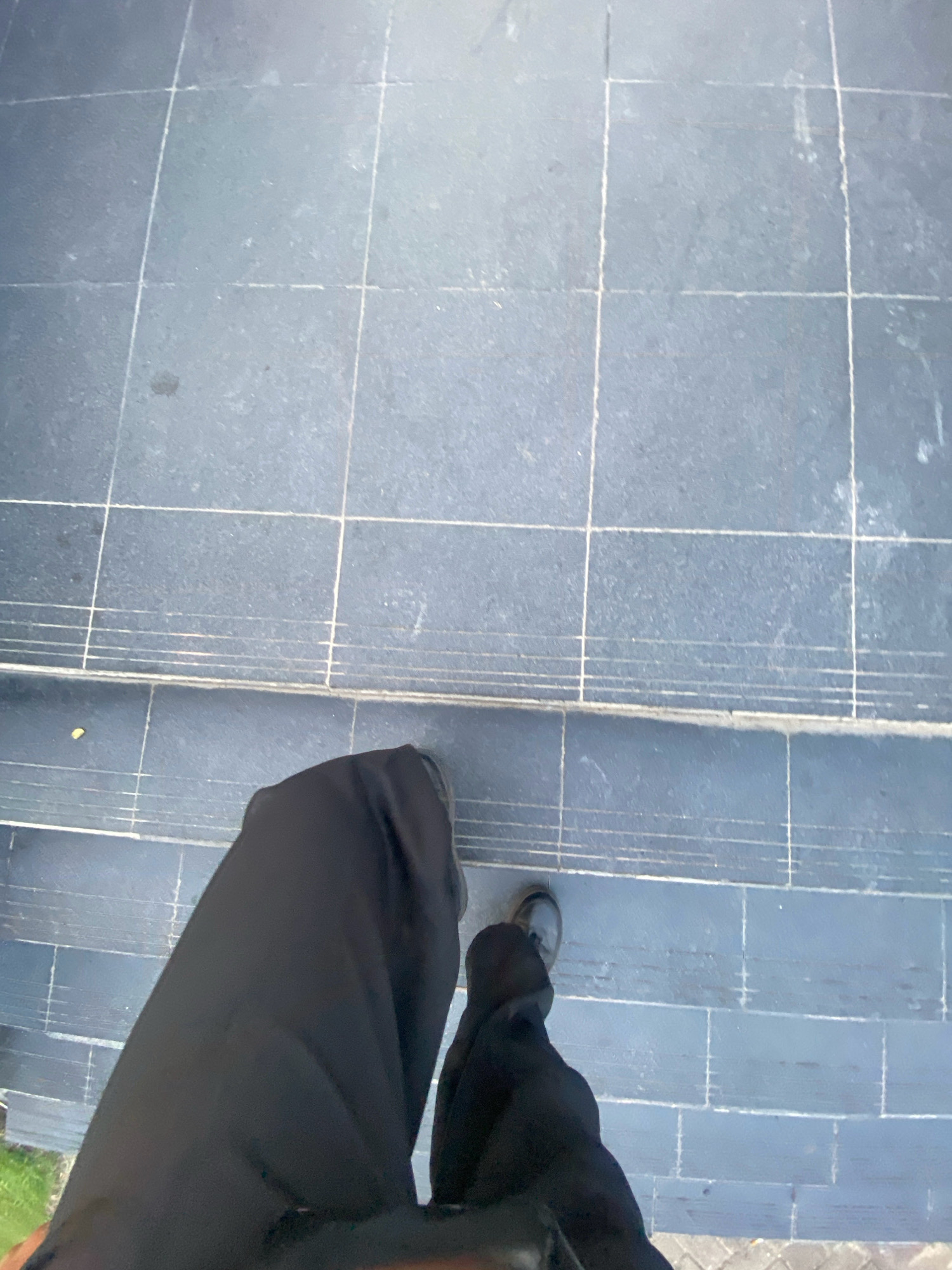The image captures a first-person perspective looking down at dark tiled stairs, likely made of stone or concrete, with visible grout lines forming a grid pattern. The tiles appear weathered and some have faint markings. In the foreground, the lower part of a person's legs and feet are visible. They are wearing black trousers and dark shoes, suggesting movement or standing on the stairs. The background shows more of the tiled surface, extending upwards and away from the viewer. A small patch of green grass is visible on the far left edge of the frame, implying an outdoor setting. The lighting suggests daytime, possibly with overcast conditions or partial shade, given the even illumination of the tiles. The environment appears to be a public outdoor area within Batam, Indonesia, possibly a walkway, entrance to a building, or a park.