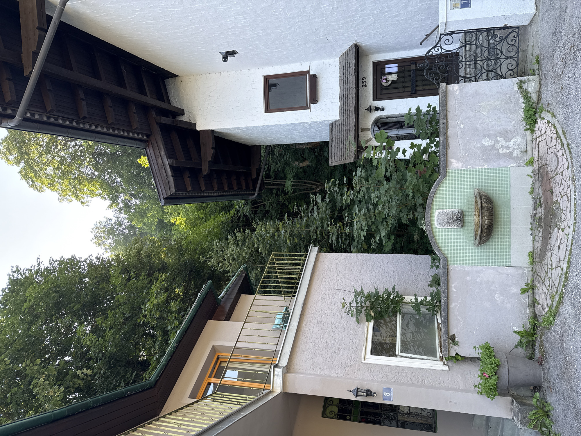 The image shows the exterior of two residential buildings in Bad Gastein, Austria, nestled against a lush green hillside. The building on the left is a multi-story structure with a light-colored stucco facade. It features a balcony with a yellow metal railing, an orange-trimmed window, and an exterior light fixture. A house number "8" is visible on a blue plaque near the entrance. The building on the right is predominantly white with a textured stucco finish. It has a dark wooden roof with exposed beams and a small pitched roof over the entrance. The entrance features a dark wooden door with a wreath and a house number "259" above it. A black wrought-iron gate with intricate scrollwork leads up a short flight of stairs. A small fountain with a decorative stone spout is integrated into a low wall separating the two properties, adorned with green mosaic tiles. The ground in front of the buildings is a mix of gravel and a circular cobblestone pattern. Overgrown greenery and trees surround the structures, indicating a natural, perhaps slightly wild, setting. The sky is clear and bright, suggesting daytime.