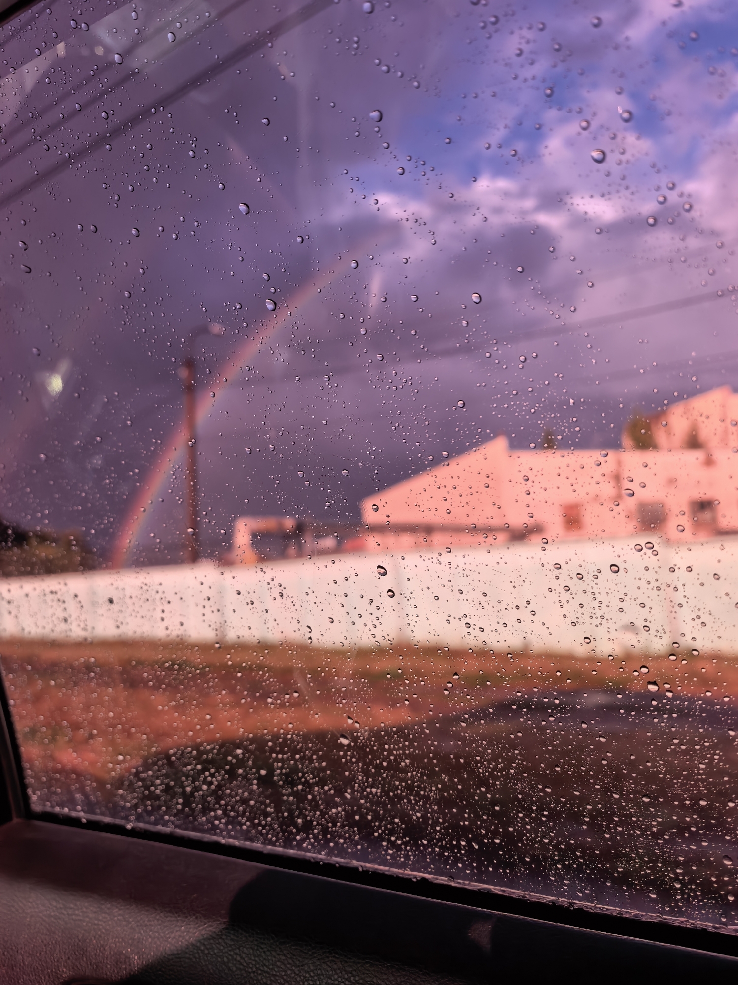 The image depicts a view through a window covered in raindrops, with a colorful sky and buildings in the background. The sky is a mix of purples and blues, with clouds illuminated by sunlight, suggesting a sunset or sunrise. A faint rainbow is visible in the upper left quadrant. In the mid-ground, there is a pink building with a white wall in front of it. The foreground shows the interior of a vehicle, with the armrest and door frame visible. The scene appears to be outdoors, possibly on the outskirts of a city, given the presence of buildings and open space. There are no people or animals visible in the image. The overall mood is atmospheric and serene, possibly hinting at a recent rain shower. The location is not specified within the image itself.