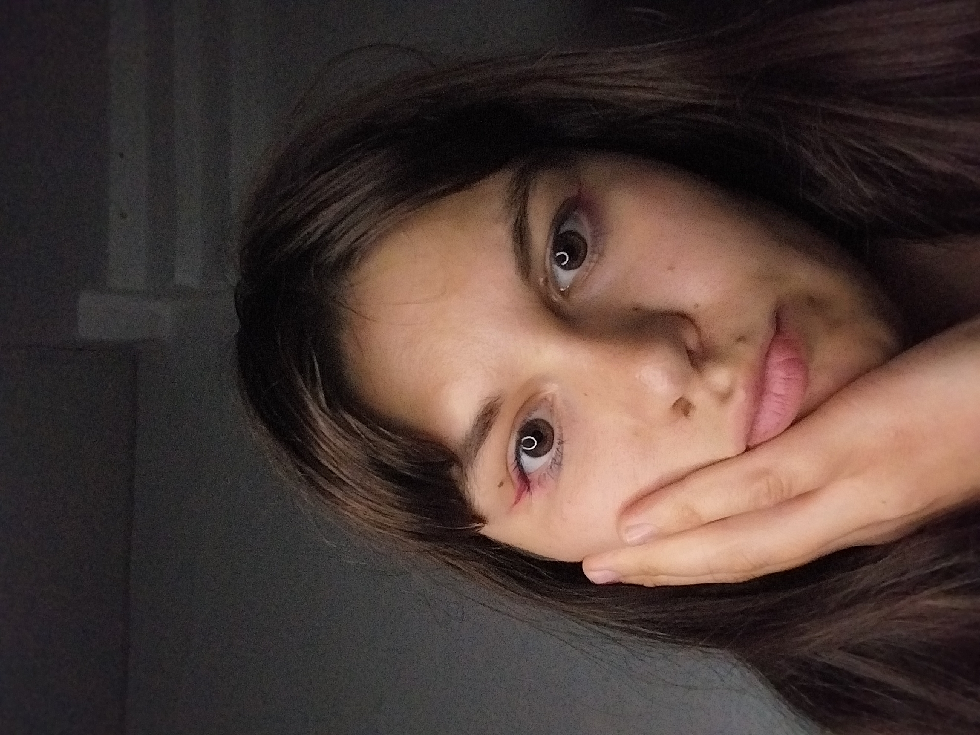 A close-up shot shows a young woman with brown hair and bangs resting her left cheek on her hand. She is wearing pink eyeliner above her eyes. The background is dark and appears to be indoors.