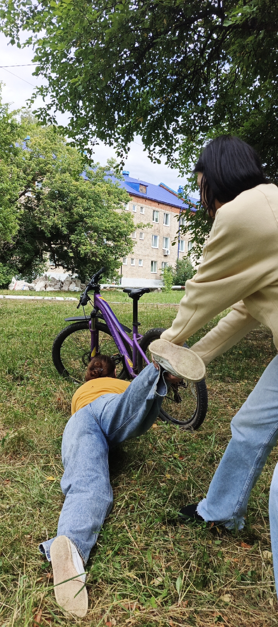 The media file shows two individuals in an outdoor setting, likely a park or grassy area with trees and a building in the background. A purple bicycle is lying on its side on the grass. One person, wearing a yellow shirt and blue jeans, is on the ground, appearing to be interacting with the bicycle. Their legs are positioned in an unusual way, with one leg raised towards the bicycle. The second person, wearing a cream-colored sweatshirt and blue jeans, is standing over the person on the ground, with one foot near the raised leg.
The building in the background is a multi-story brick structure with blue roofs, suggesting an urban or suburban environment. The presence of trees with green leaves indicates it is likely spring or summer. The lighting suggests it is daytime, possibly overcast given the soft shadows. The overall mood appears playful or perhaps a staged comedic moment, with the awkward positions of the individuals and the fallen bicycle. No visible text is present in the image. The location context provided is Chaikovsky, Russia.